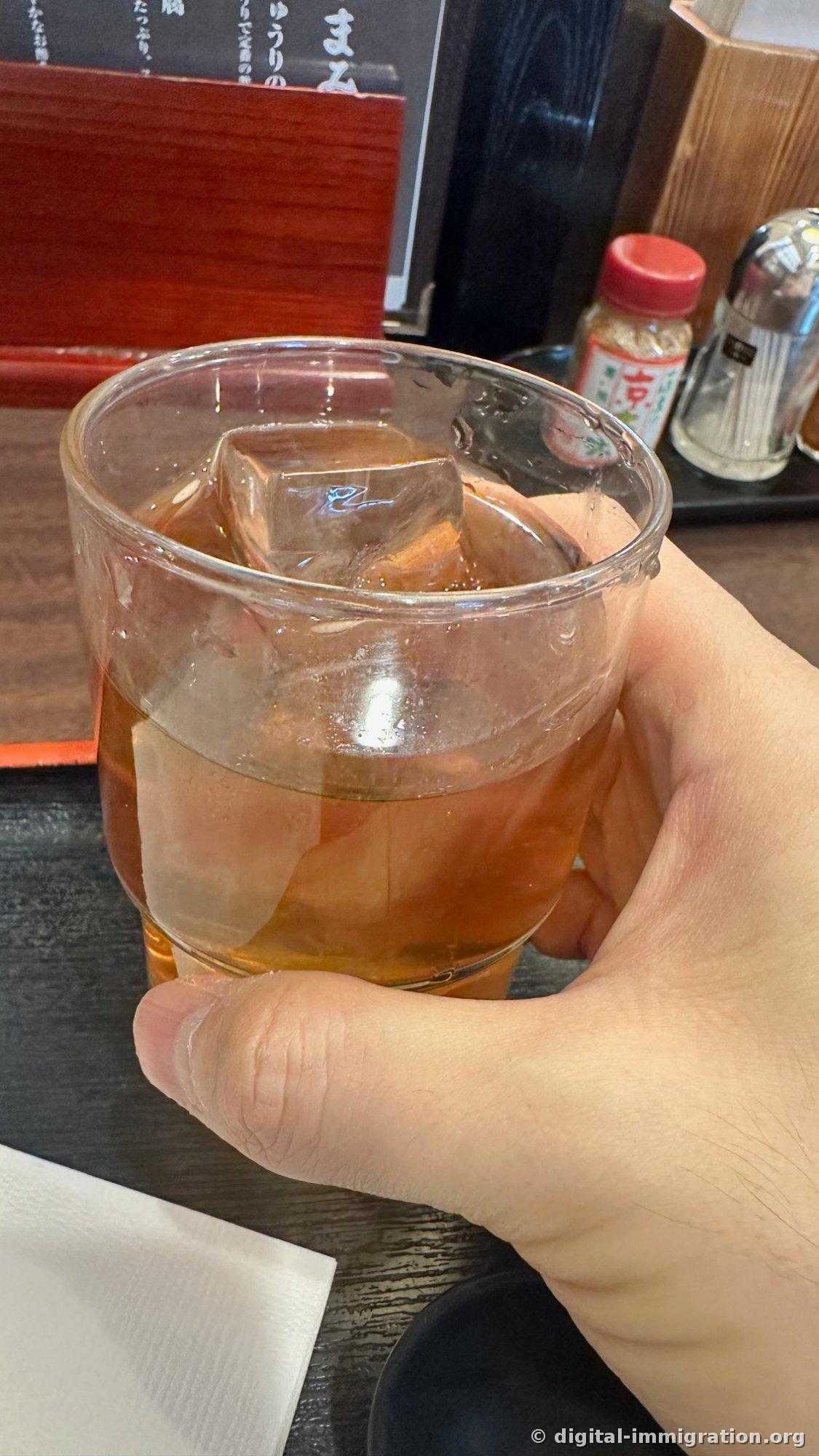 Un verre tenu dans la main avec de gros glaçons cubiques et du thé brun, et à l'arrière la bouteille de shichimi de Kyoto avec un bouchon rouge et un poivrier en argent
