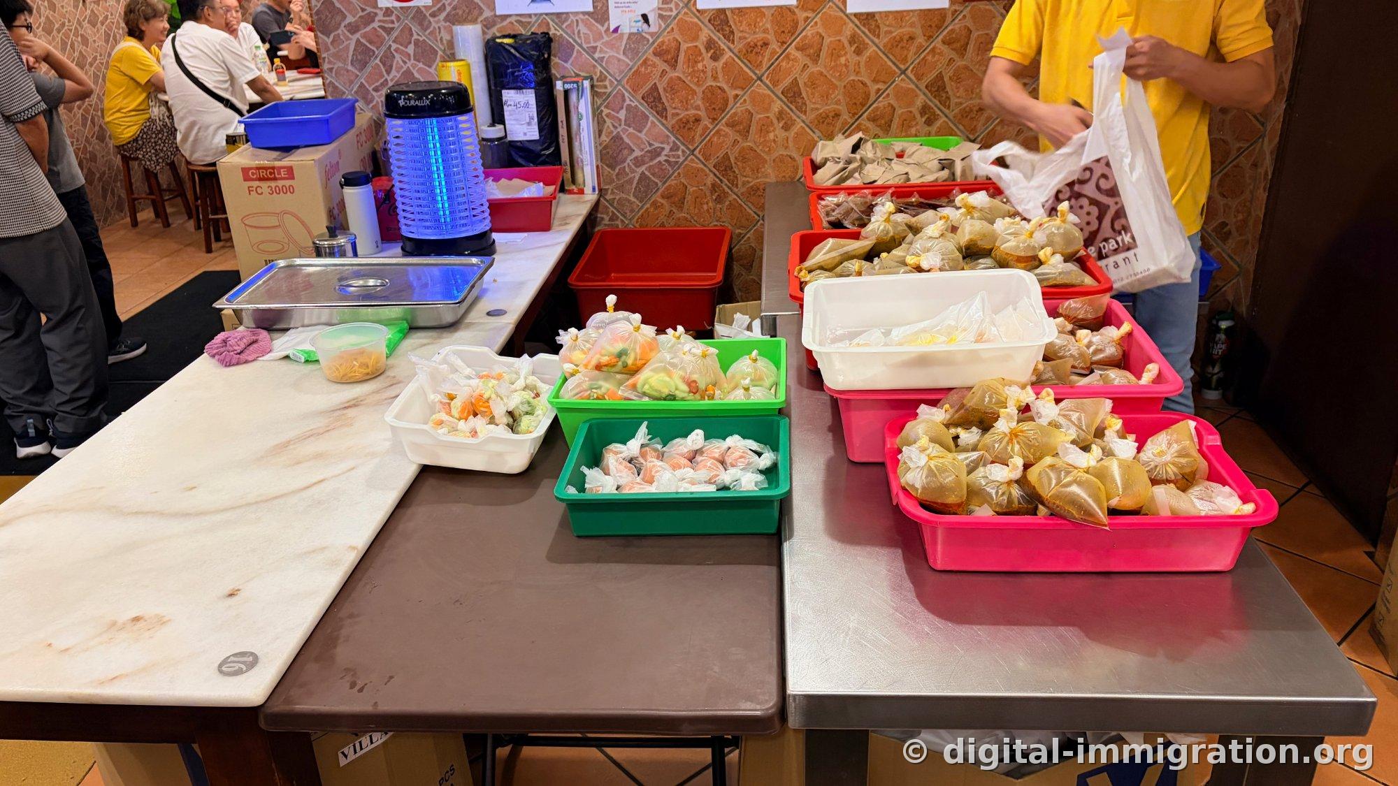 Plastic trays of various colors piled with plastic bags filled with sambal and side dishes sit on a marble counter, while a staff member in a yellow uniform packs them into white plastic bags