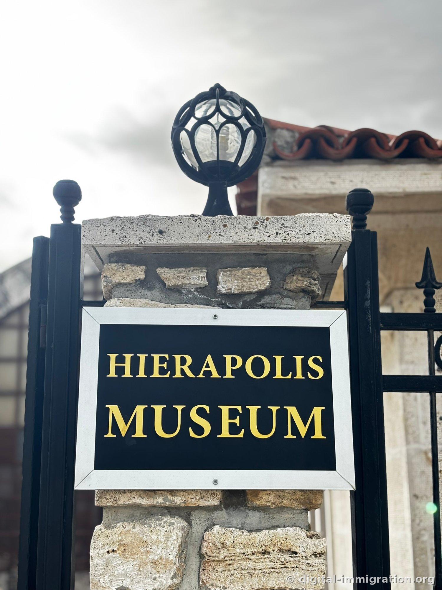 Entrance sign reading 'HIERAPOLIS MUSEUM' in gold letters on a black background, mounted on a metal gate pillar with a round lamp fixture above