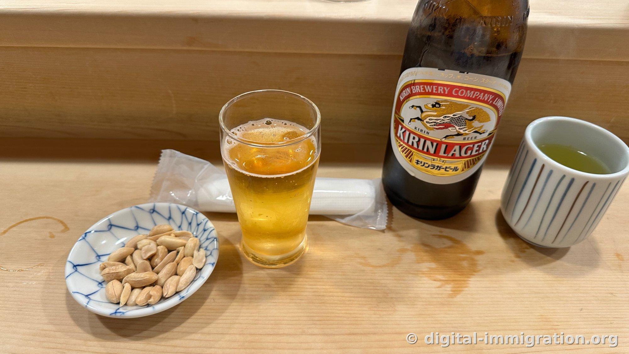 On the counter: a small dish with blue pattern holding peanuts, a glass of beer, a bottle of Kirin Lager, and green tea in a striped teacup