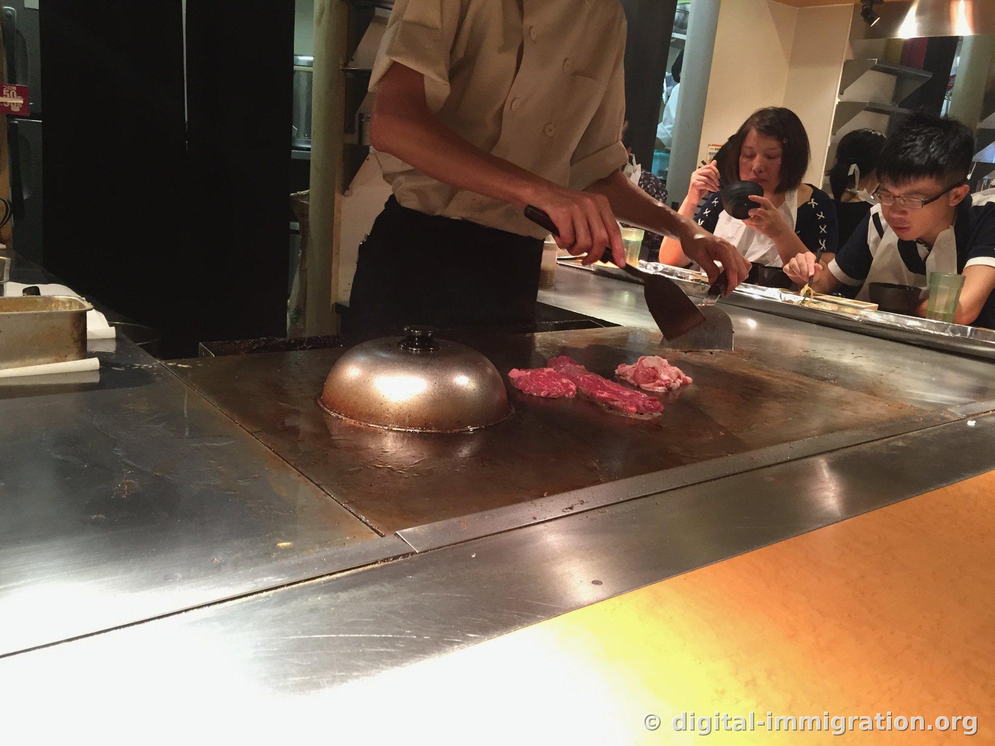 Un cuisinier en tenue blanche fait cuire du boeuf rouge et des tripes blanches couvertes de gras sur une grande teppan avec une spatule, un couvercle métallique circulaire posé à côté. Les clients au comptoir en face sont en train de manger