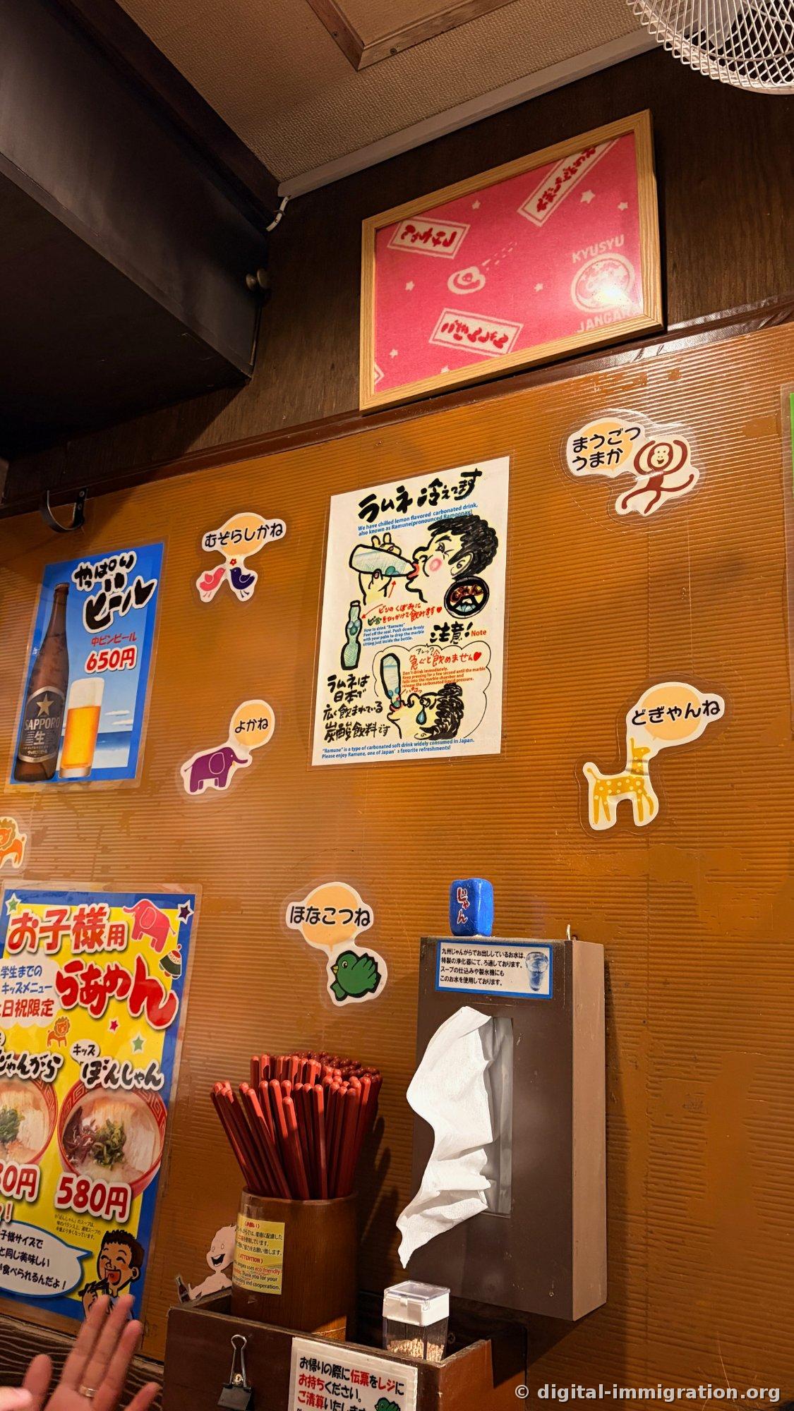 Kyushu dialect speech bubble stickers and ramune drinking instruction poster on brown wall, Sapporo beer 650 yen poster, children's ramen 580 yen poster, red chopstick dispenser and napkin box