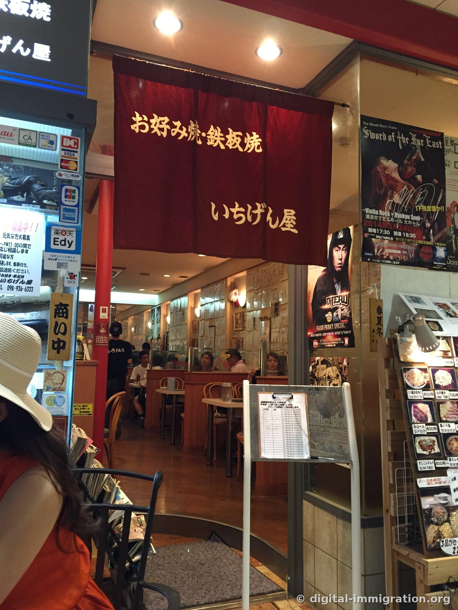 Shop entrance with a red noren and 'お好み焼・鉄板焼 いちげん屋' written in gold letters, the waiting list clipboard in front, and the menu photo board on the right
