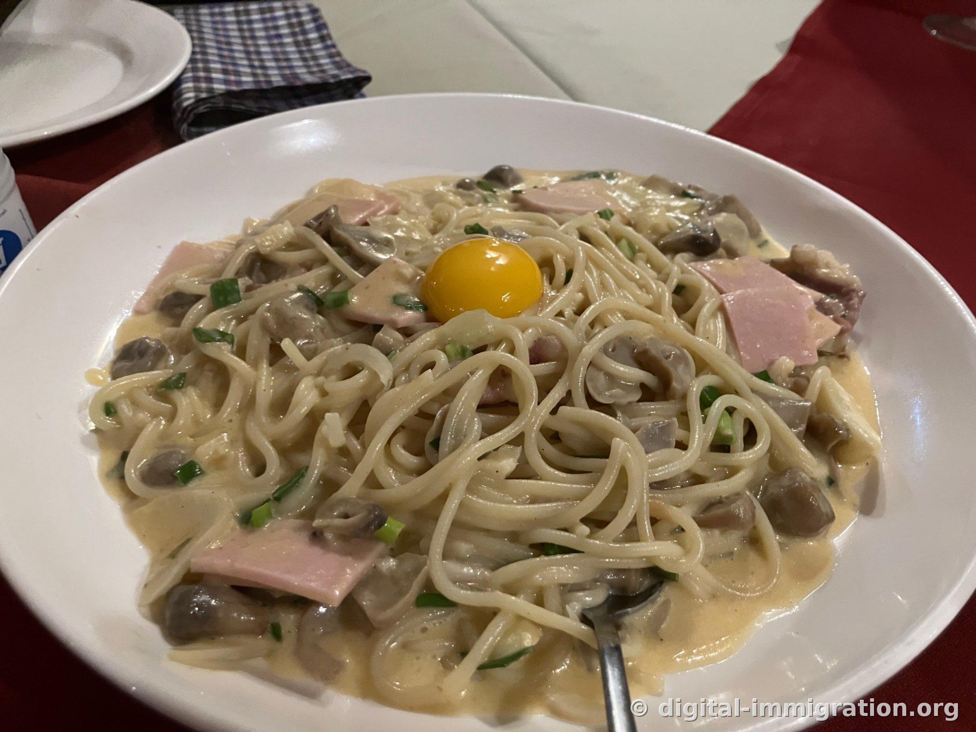 Creamy spaghetti carbonara in a white bowl, mixed with ham, mushroom pieces, and chopped scallions, with a raw yolk on top