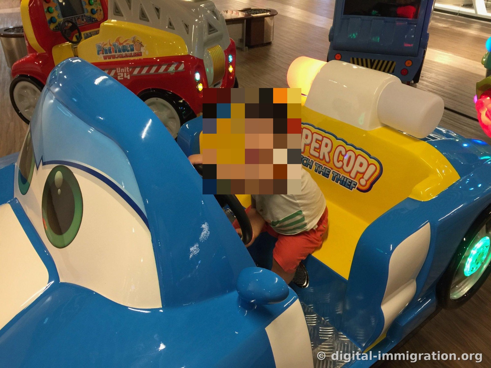 Un manège pour enfants à pièces dans un passage du centre commercial. Un enfant est assis dans la voiture de police bleue « SUPER COP » en tenant le volant, avec un camion de pompiers rouge et un bus bleu visibles derrière