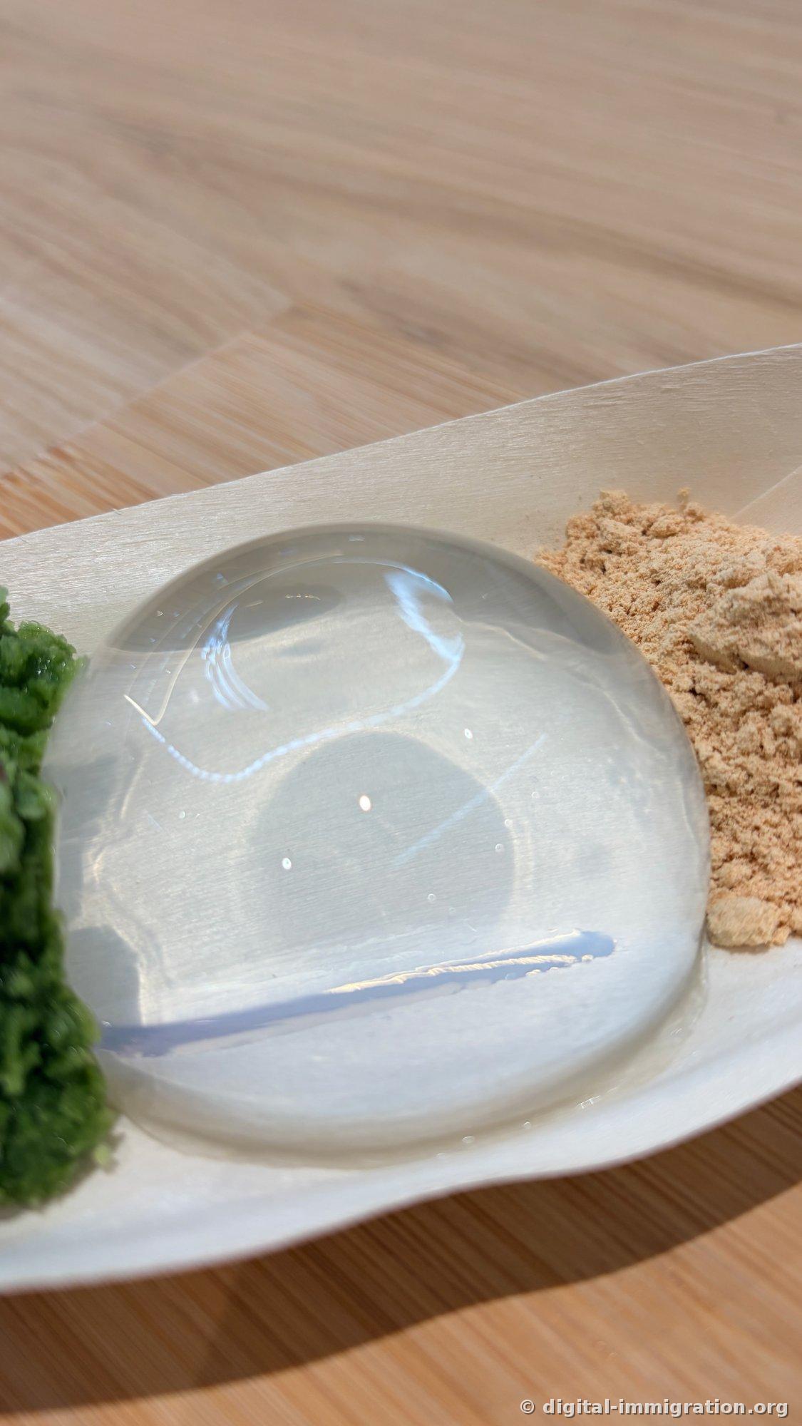 Close-up photo of transparent water drop mochi on a wooden plate, with ceiling lighting reflected on its surface, green matcha powder on the left and soybean powder on the right