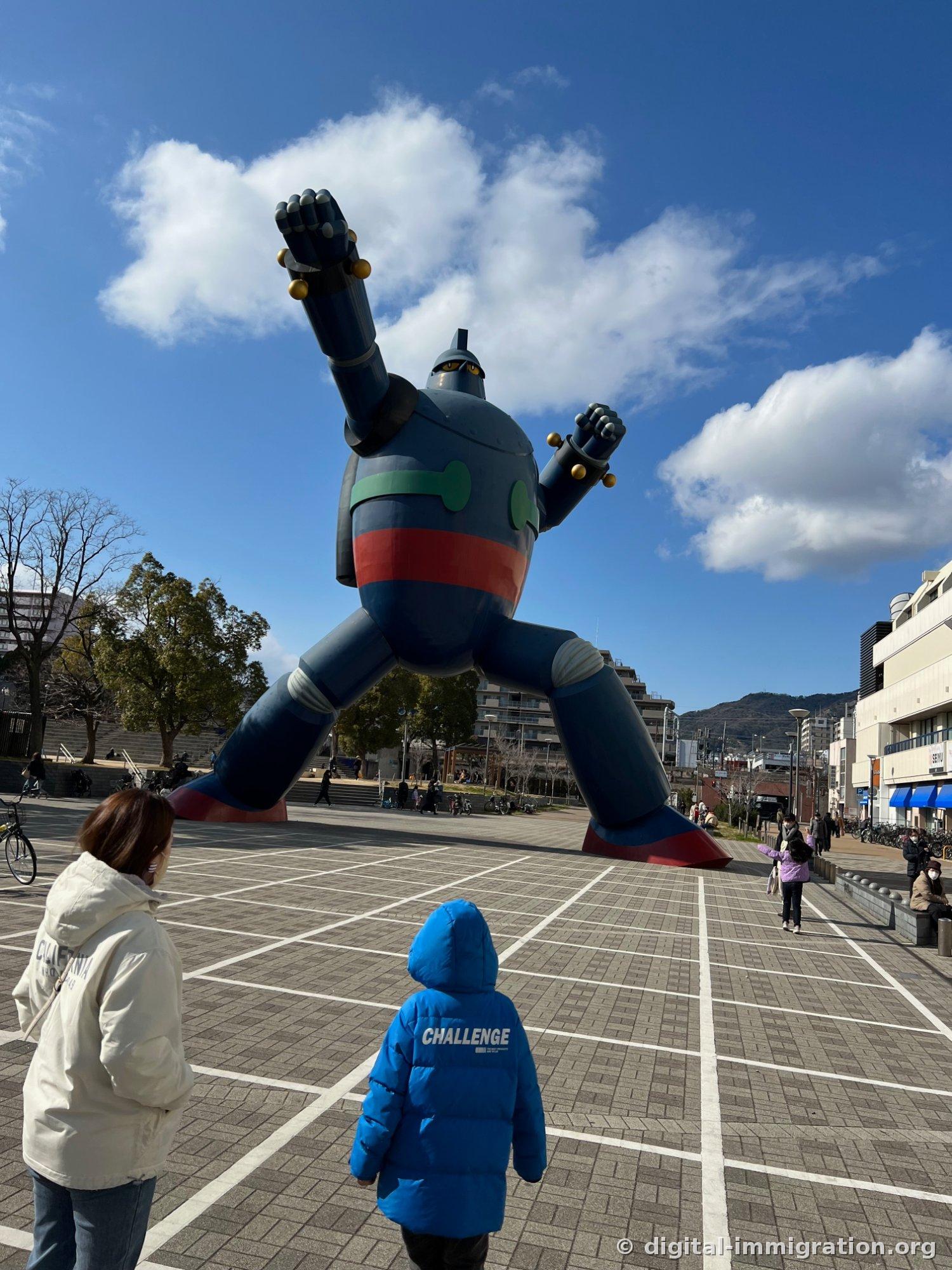 Sous un ciel bleu, le immense monument Tetsujin 28 avec le bras droit levé, regardé par un enfant et un adulte sur la place