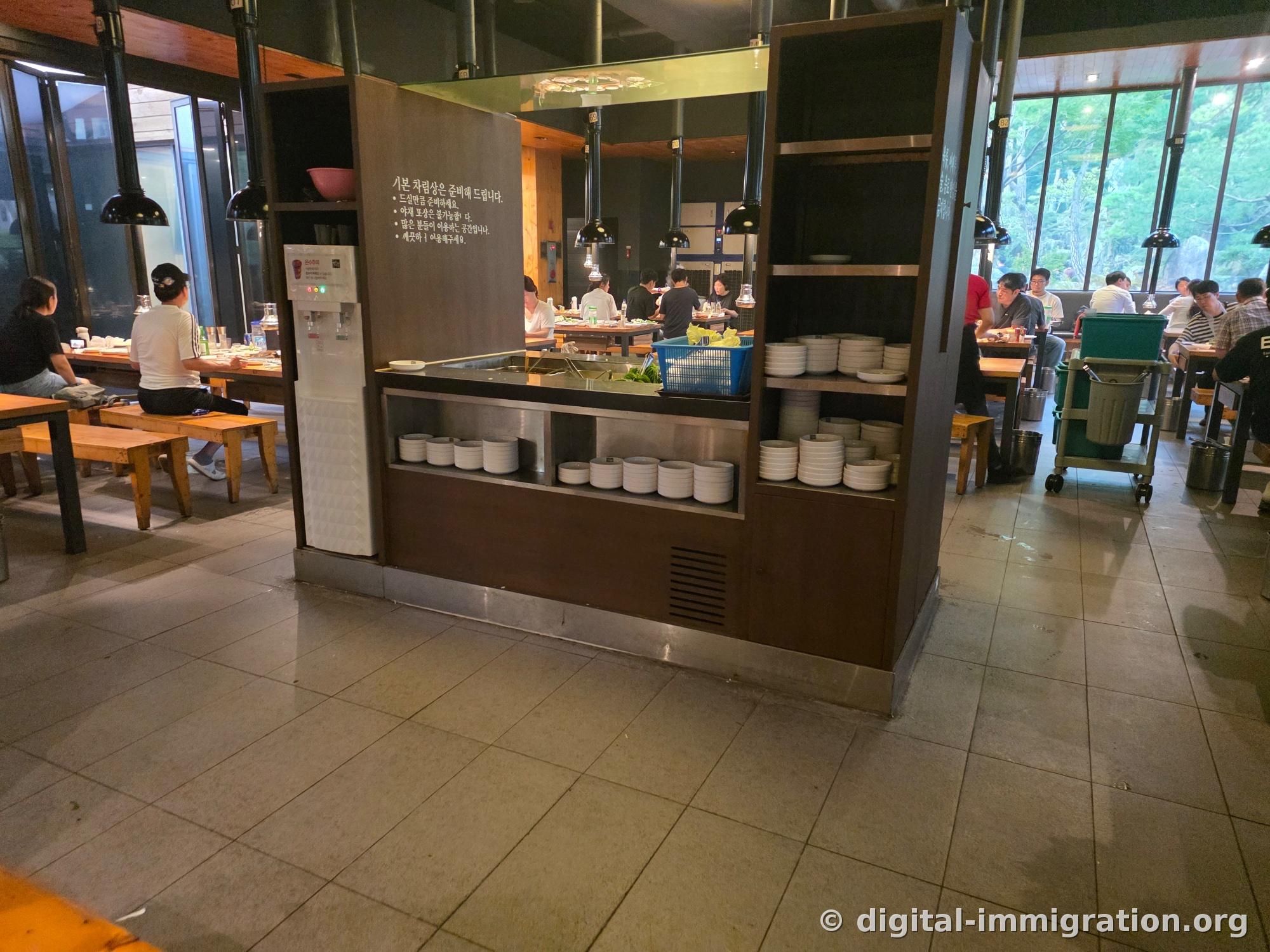 Le bar libre-service au centre de la salle de la Ferme Galilée avec des instructions murales comme « Les accompagnements de base sont préparés », « Servez-vous selon vos besoins », et les assiettes blanches empilées sur les étagères