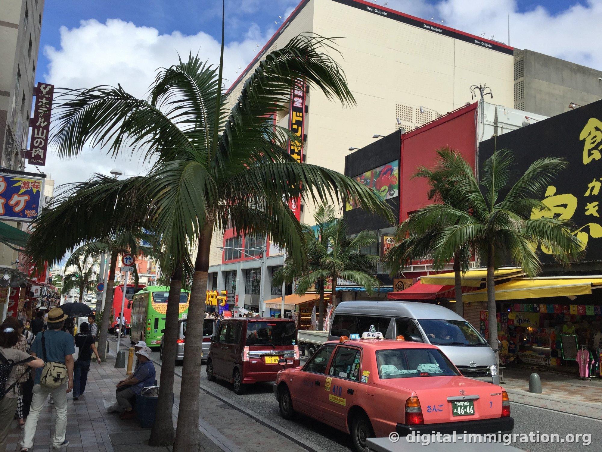 La rue Kokusai-dori de Naha bordée de palmiers, avec le bâtiment Donki Hote (ドン・キホーテ) à droite et des magasins aux auvents jaunes, un taxi rose Okinawa garé sur la route