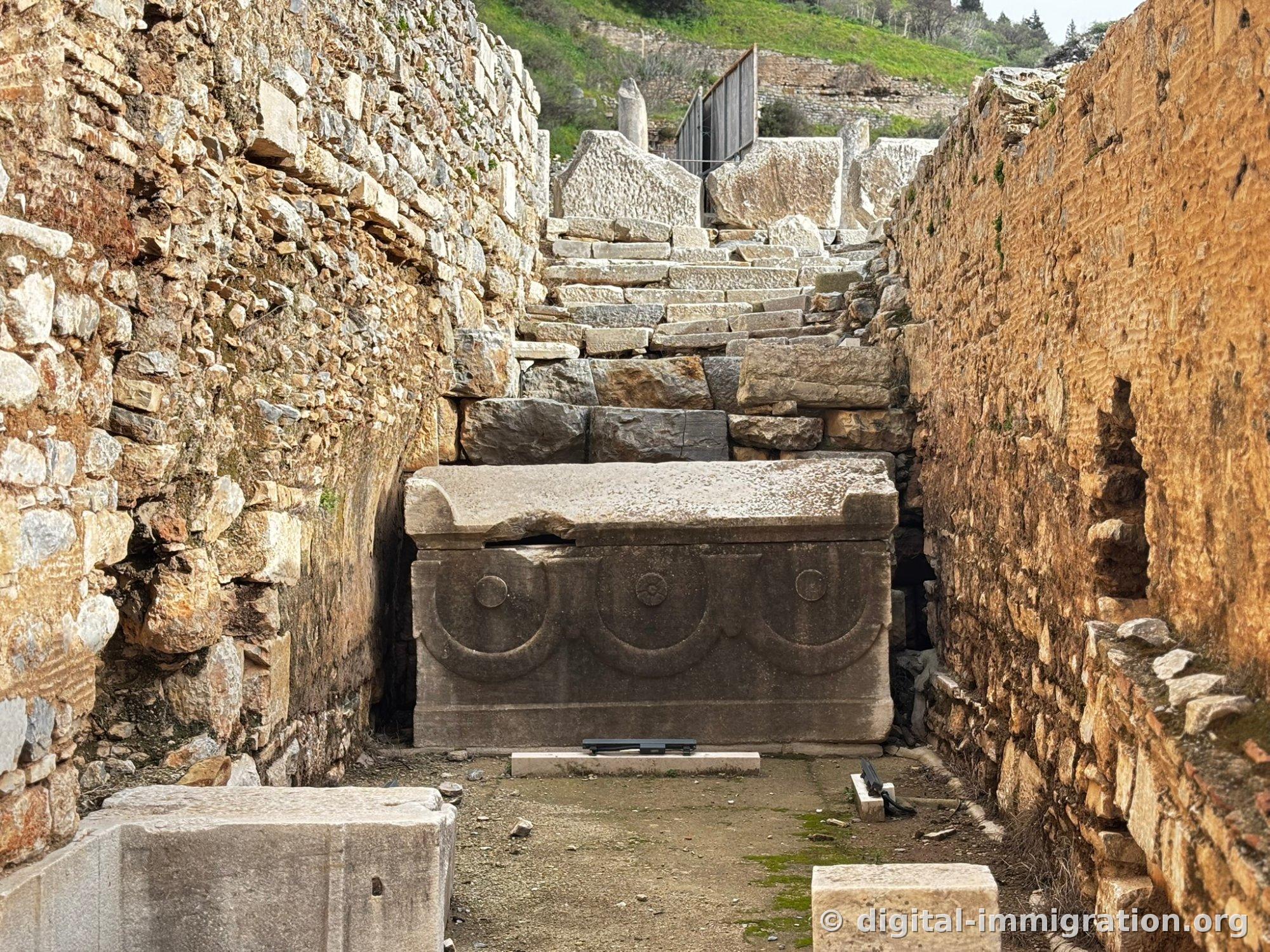 A marble sarcophagus with garland relief carvings sits at the end of a narrow passage between stone walls, with stone steps continuing beyond