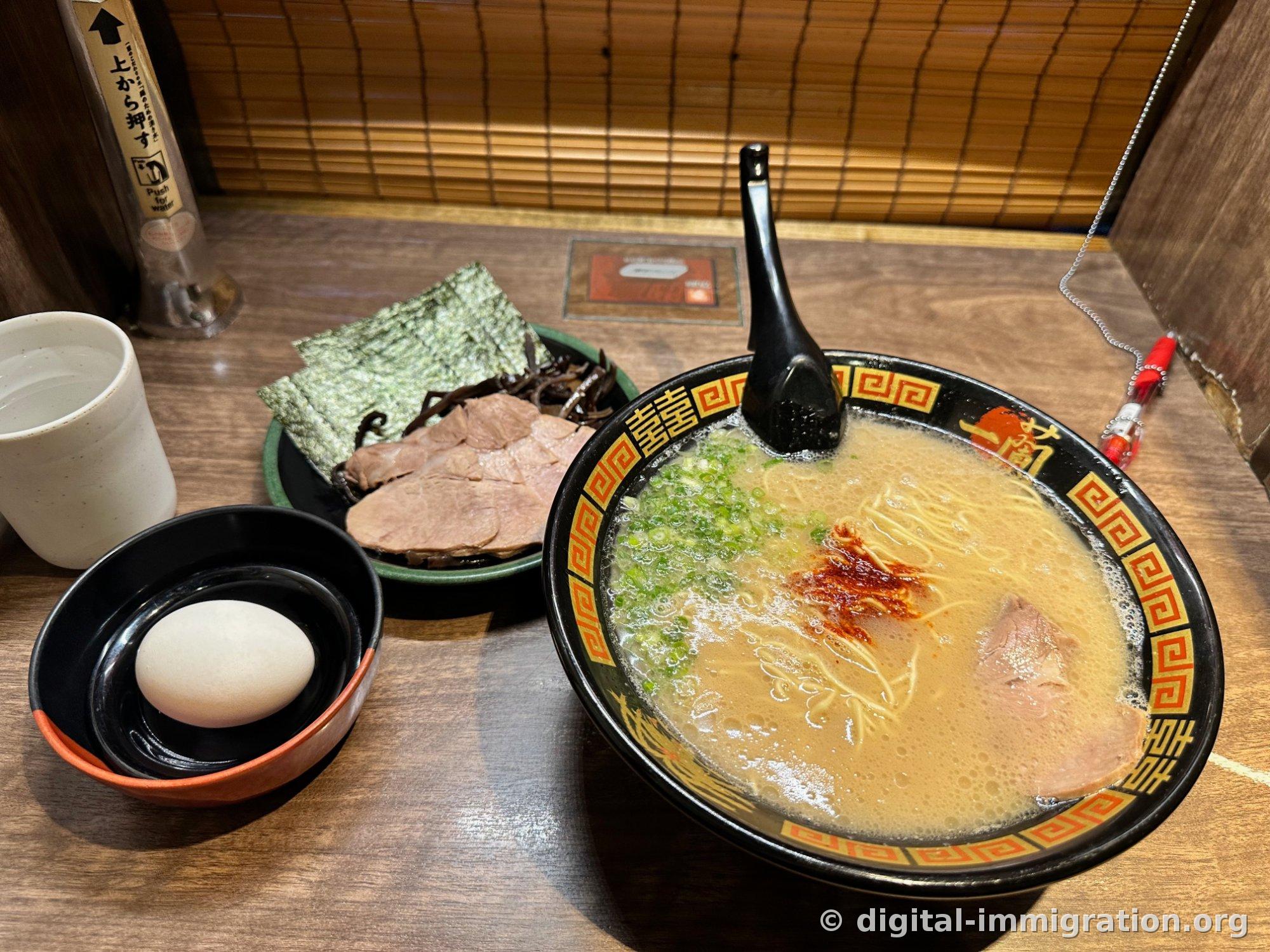 Mise en place complète d'une portion : un bol de ramen, une assiette avec char siu et kikurage, un petit bol avec l'œuf, une tasse d'eau et la cuillère noire, tous alignés sur le comptoir