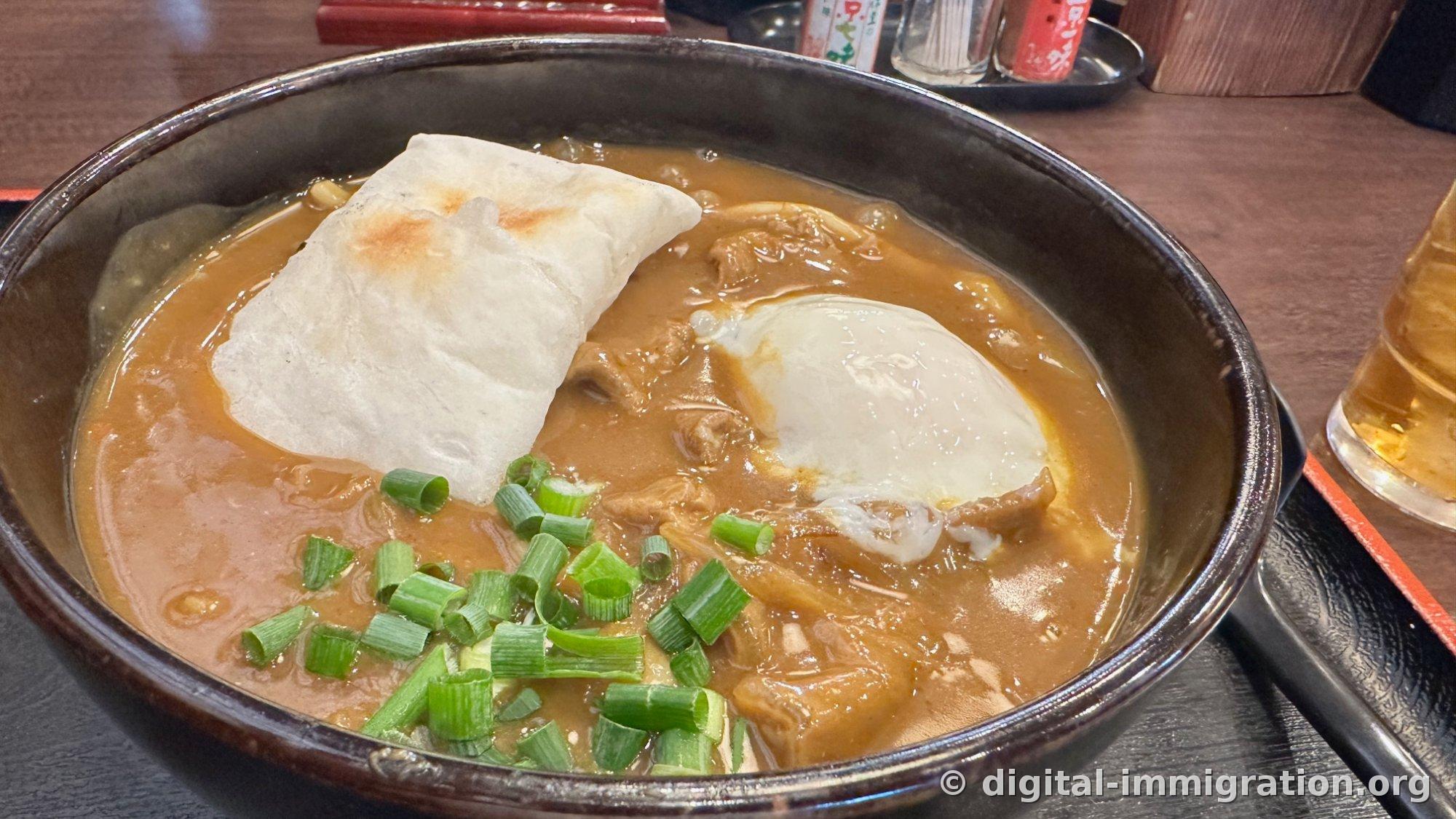 Curry udon d'un brun épais dans un bol noir. Sur la gauche, une grande feuille d'aburaage grillée, sur la droite un œuf avec le blanc solidifié, au centre de la viande fine et des oignons verts hachés