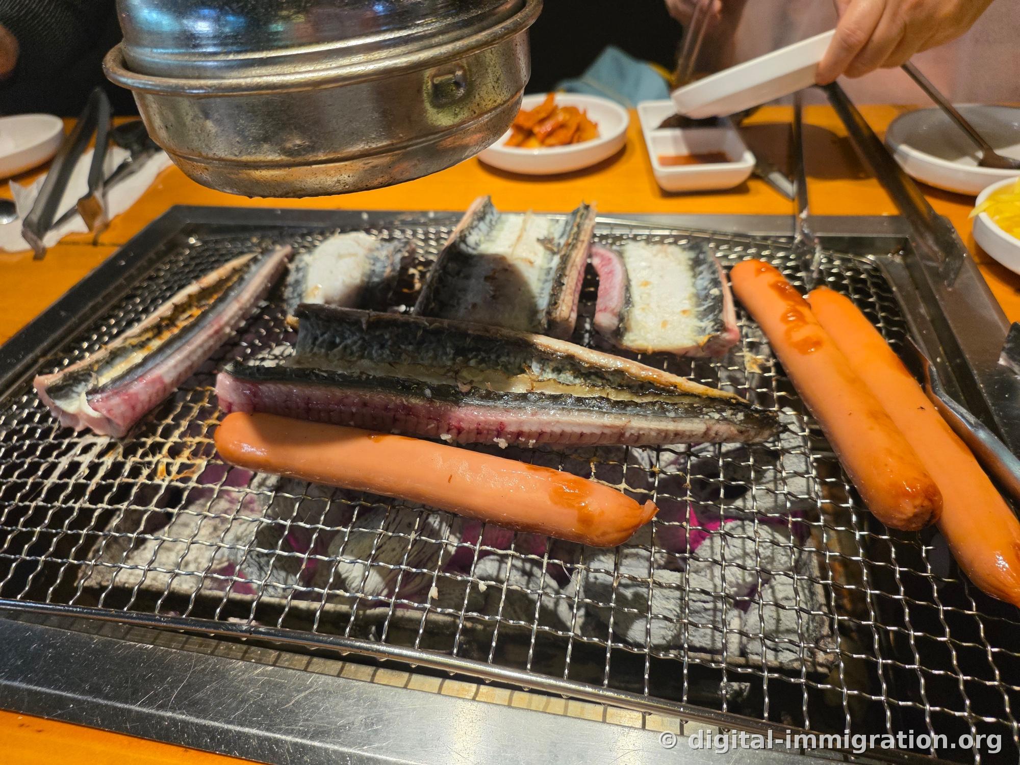 Sur la grille du feu de charbon, plusieurs morceaux d'anguille nettoyée avec la peau noire et la chair rose qui cuisent, avec deux saucisses qui cuisent aussi à côté