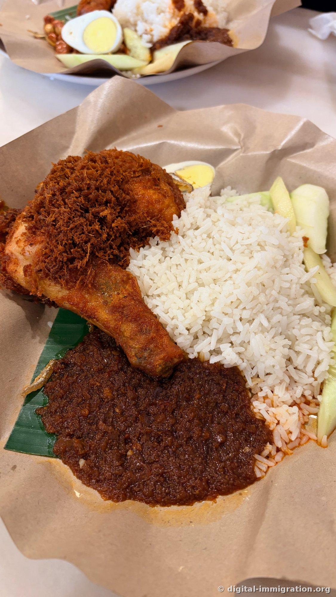 On brown paper, white coconut rice, cucumber, soft-boiled egg, dark brown sambal on a banana leaf, and a fried chicken leg thickly coated with spiced fried crumbs sit on a plate of nasi lemak