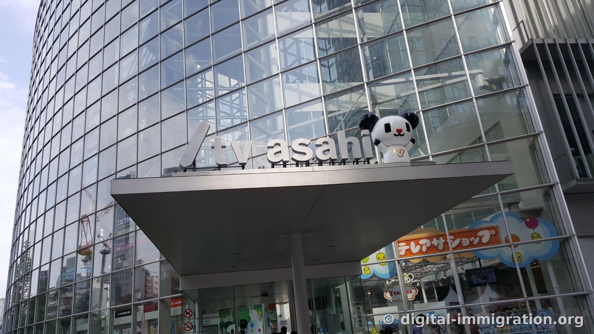 Panneau blanc 'tv asahi' fixé sur la façade du bâtiment à murs-rideaux vitrés avec une figurine de panda blanc assise à côté, et l'enseigne TeléAsaShop visible en bas à droite