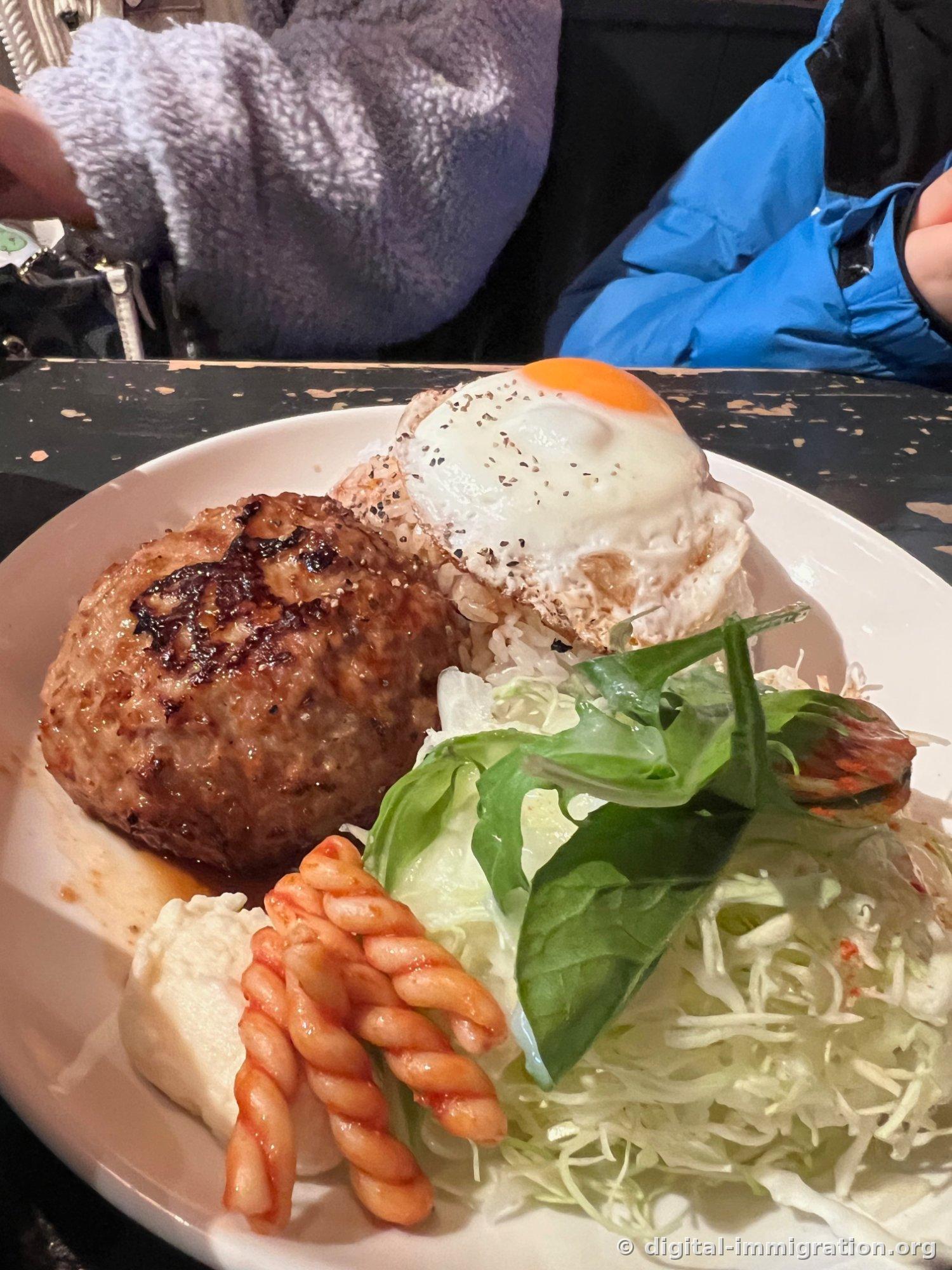Set de steak haché servi dans une assiette blanche avec un bifteck haché, du riz garni d'un œuf frit semi-cuit, du chou râpé, de la laitue, une salade de pommes de terre et des pâtes enrobées de ketchup