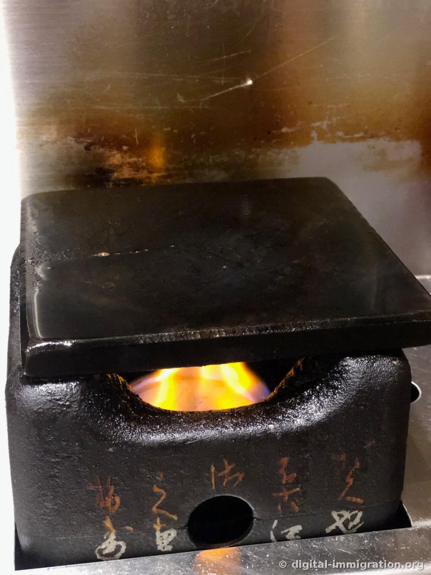 Pierre carrée noire posée sur un support en fonte. Les flammes de combustible solide orange brûlent sous le support, et des lettres rouges et blanches sont écrites sur le côté du support