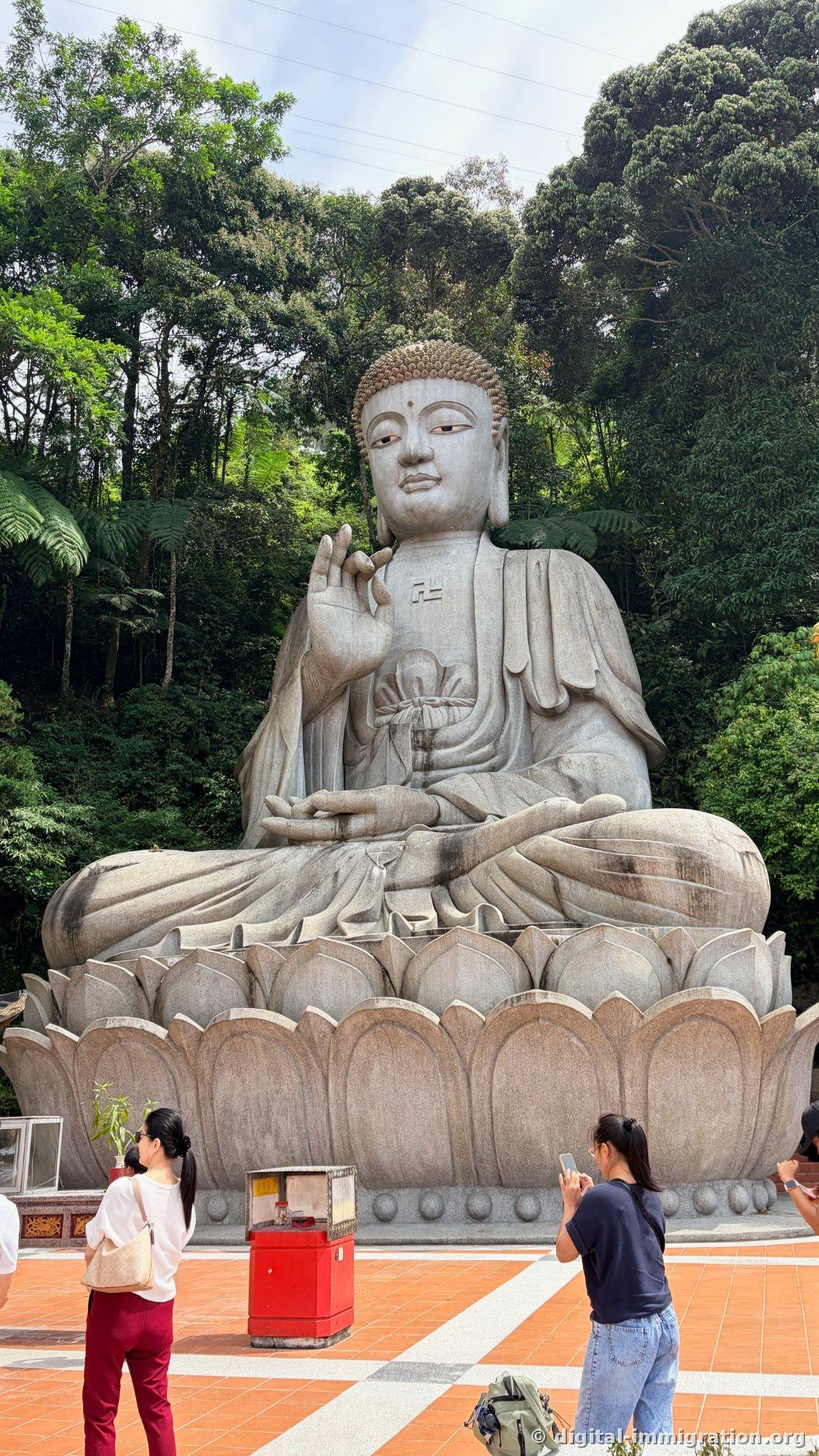 Vue rapprochée de la gigantesque statue assise de Bouddha en pierre, levant légèrement la main, avec le caractère Sanskrit sur la poitrine et le siège de lotus visible, des touristes prenant des photos en bas