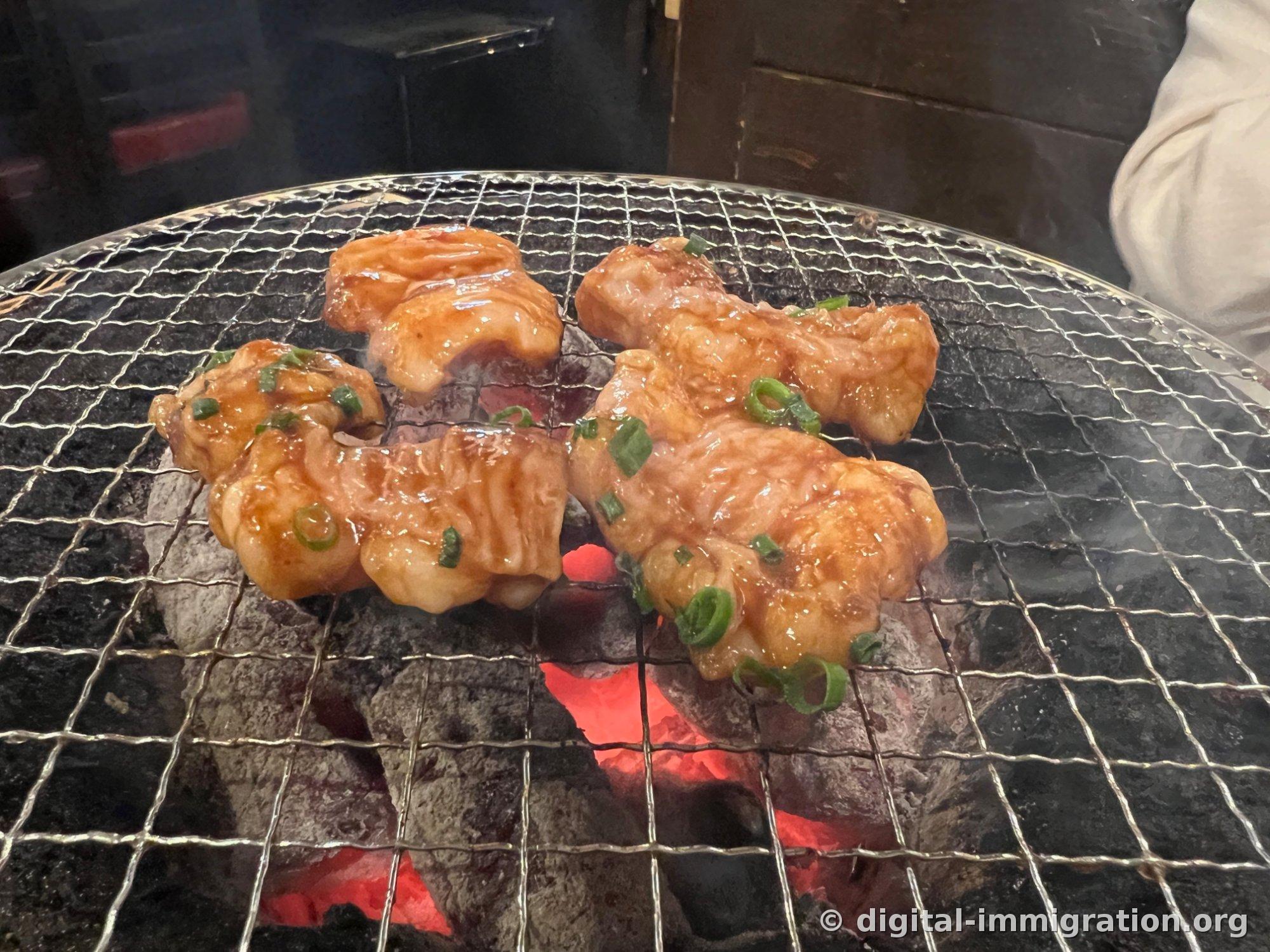 炭火網の上で火が通っているタレ大腸がいくつか。表面にツヤが出ていて小ネギが乗っていて、下の炭が赤く熱くなっている