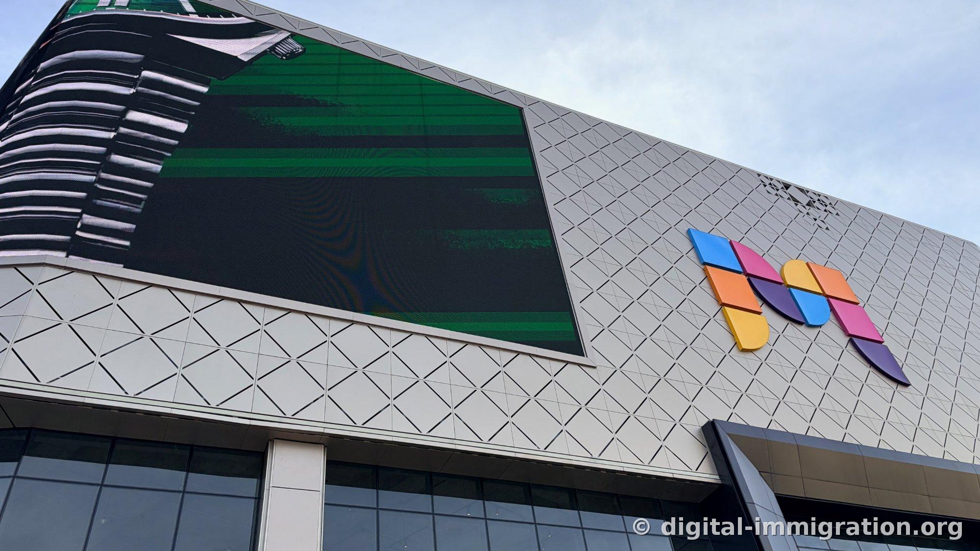 Mall façade viewed from below, triangular pattern panels and colorful logo pieces, large LED screen on the side