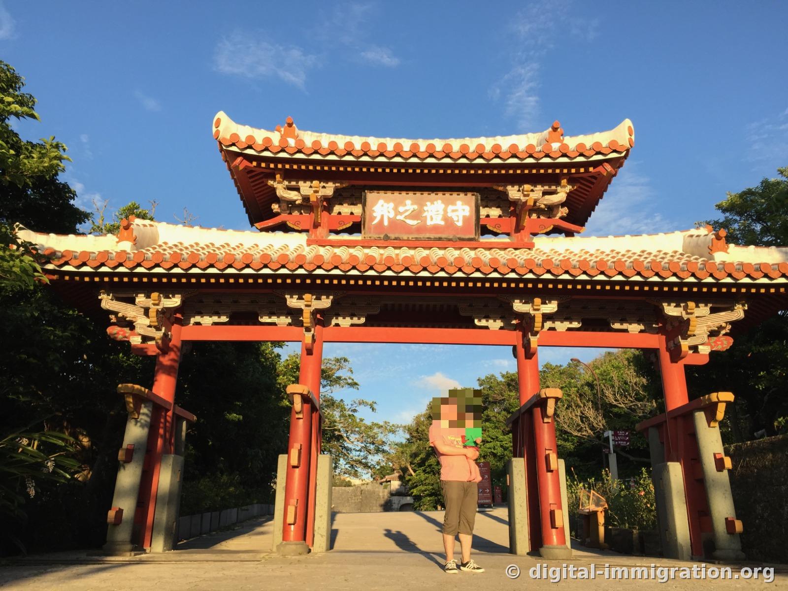 Holding a child under the vermillion Shureimon with the plaque reading '守禮之邦', blue sky visible behind