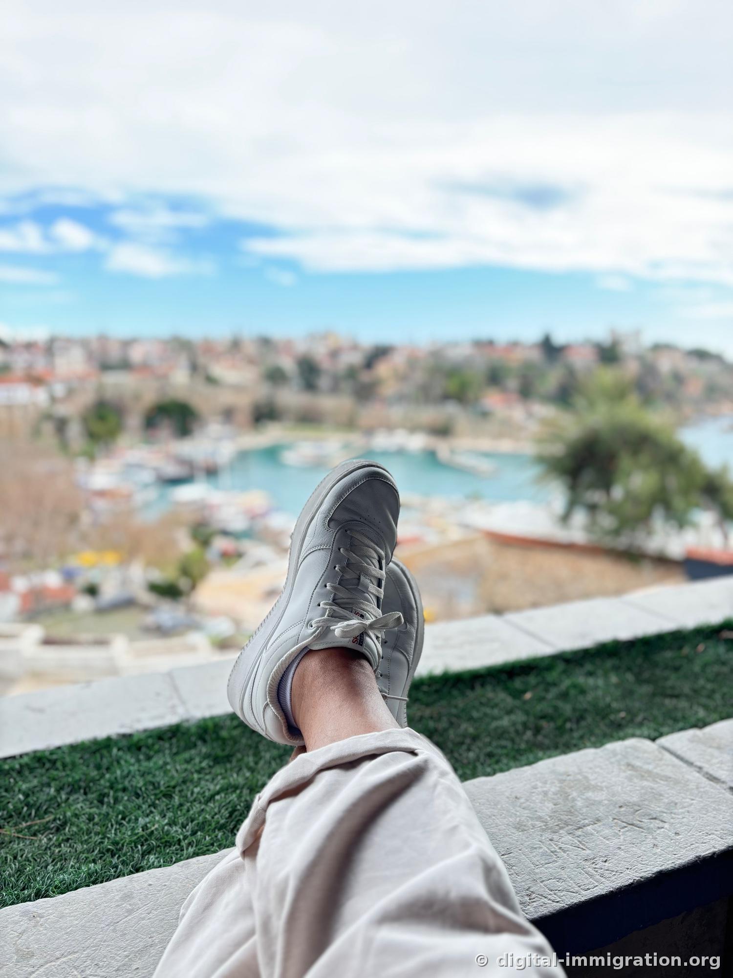 First-person view with white sneakers propped up on the cliff-top café railing, harbor below framing the jade-colored Kaleici yacht harbor out of focus