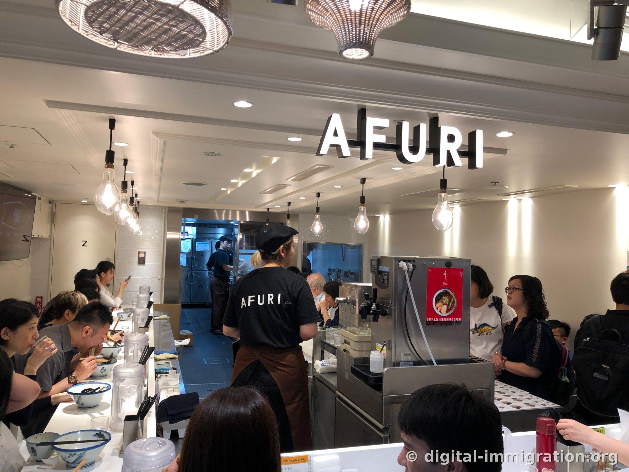 Employee in black AFURI logo t-shirt standing behind the counter, with customers eating ramen at counter seating