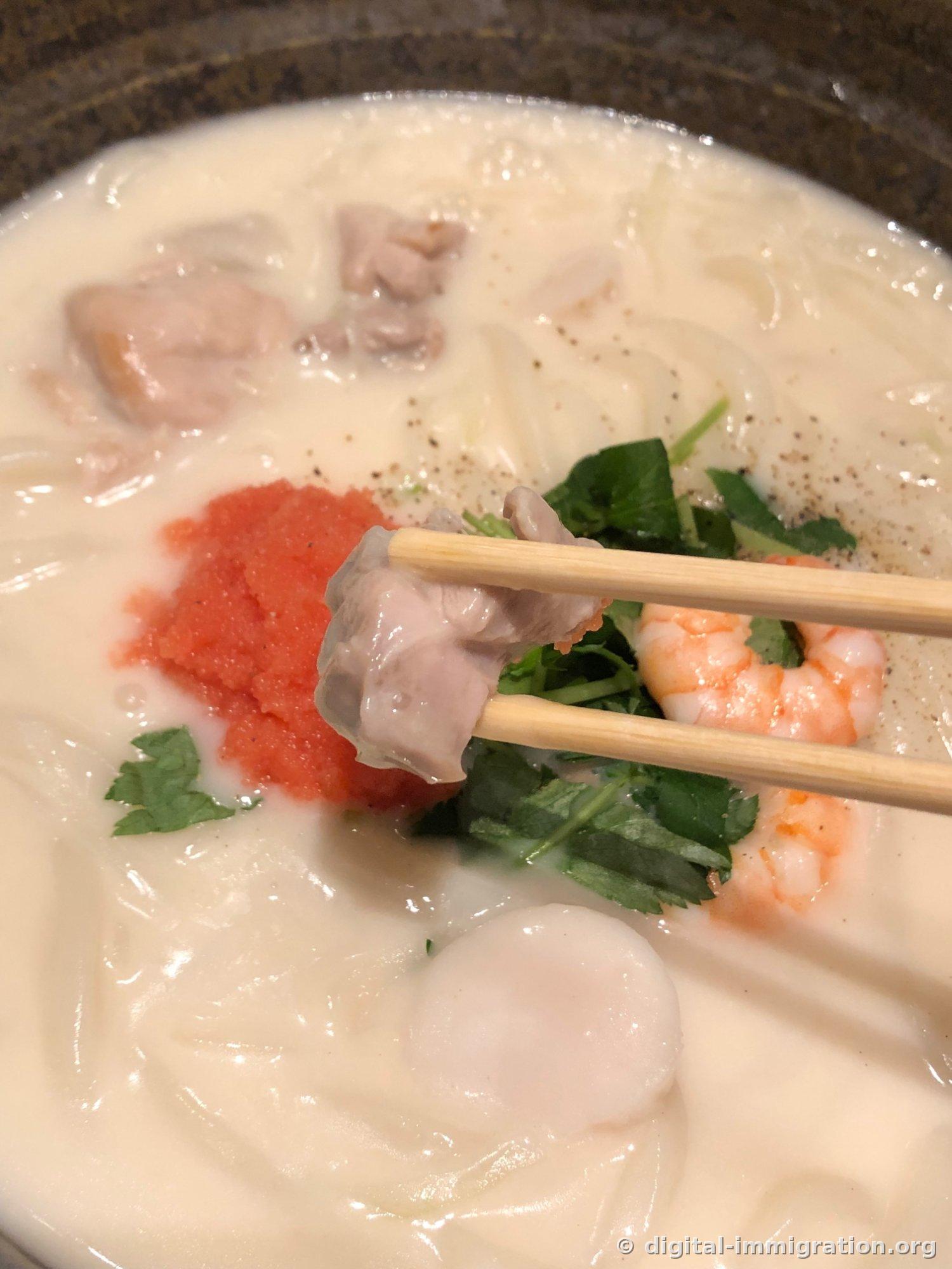 A chunk of chicken with skin gripped by chopsticks, with cod roe and scallop visible below