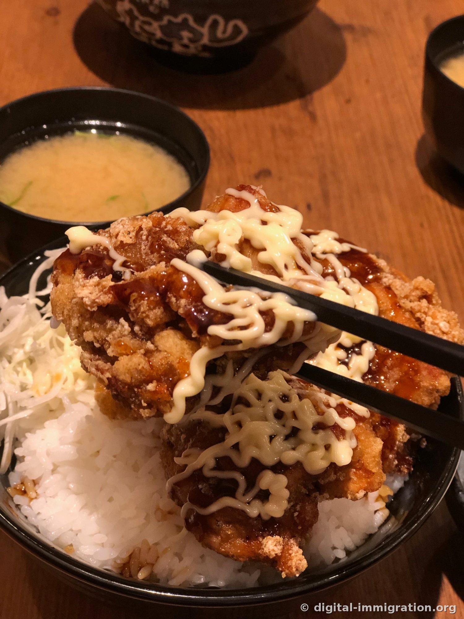 Un morceau de poulet frit garni de sauce et de mayonnaise soulevé avec des baguettes, avec deux bols de soupe miso à l'arrière-plan
