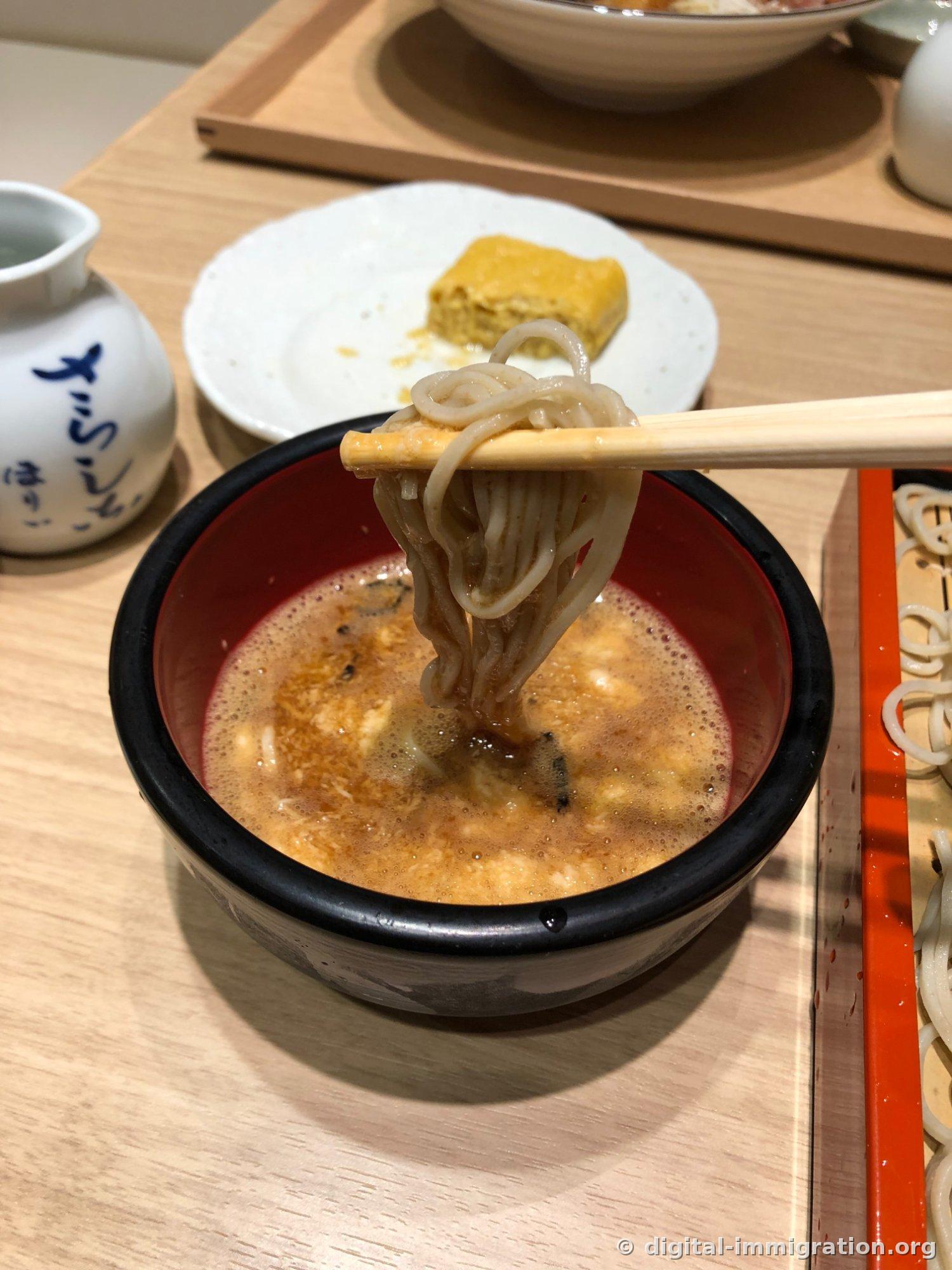 Lifting soba with chopsticks beside a white porcelain cup with 'さらしなほりい' written on it, with remaining tamagoyaki on the plate behind