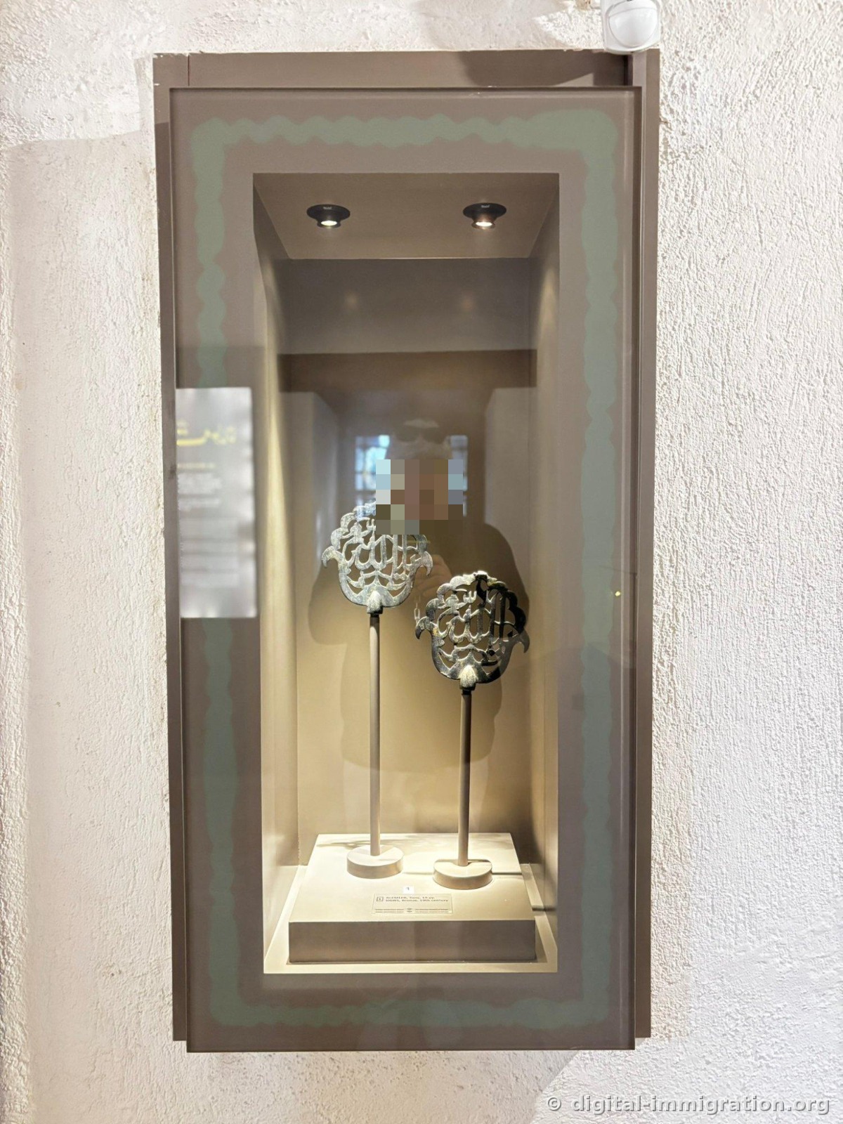 Dans une niche vitrine murale, deux finiales en bronze du 19e siècle sous projecteur, avec le reflet du photographe superposé dans le verre