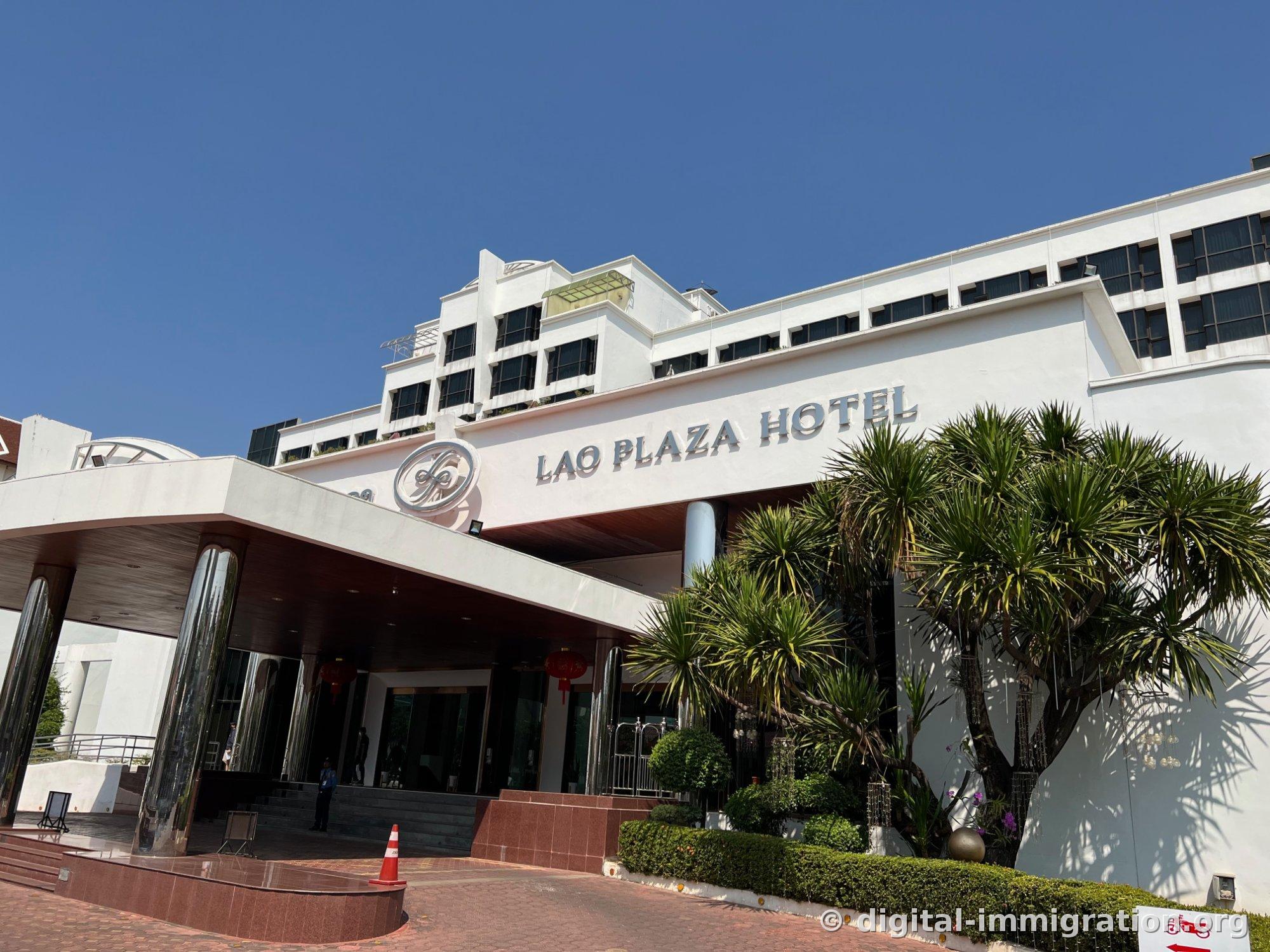 Façade principale de l'hôtel avec auvent en verre et panneau argenté LAO PLAZA HOTEL sous un ciel bleu avec des palmiers yucca