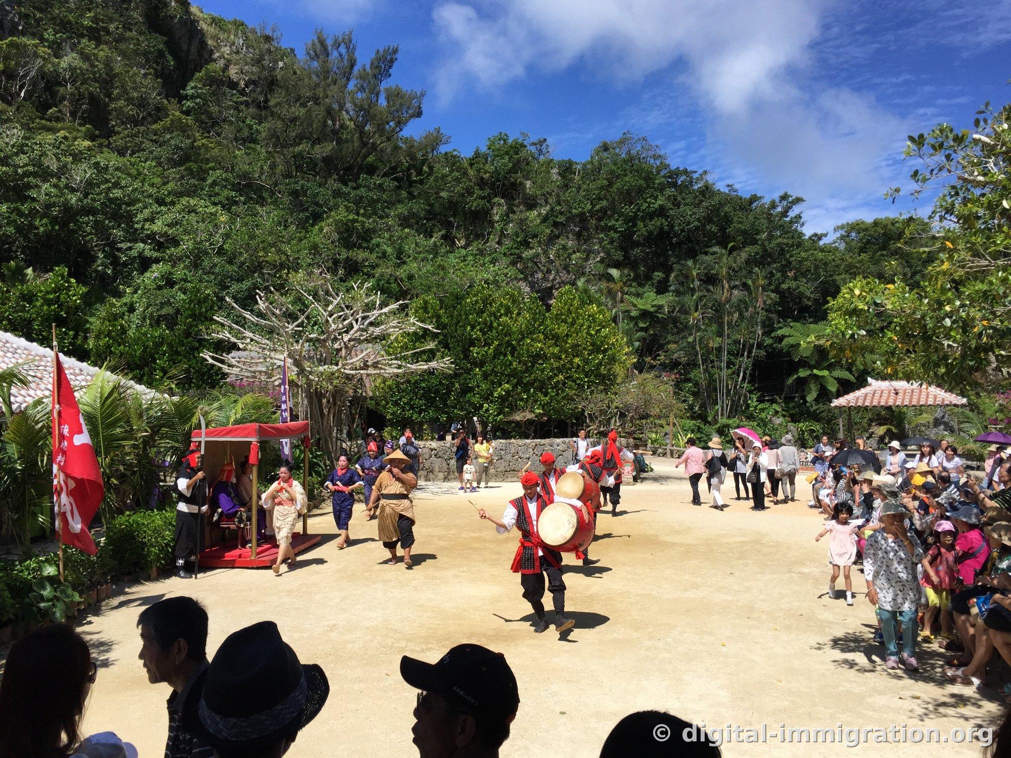 Au centre de la cour du Ryukyu Mura, une procession avec un palanquin rouge et des drapeaux, tandis qu'un musicien jouant du tambour court en première ligne, entouré de spectateurs