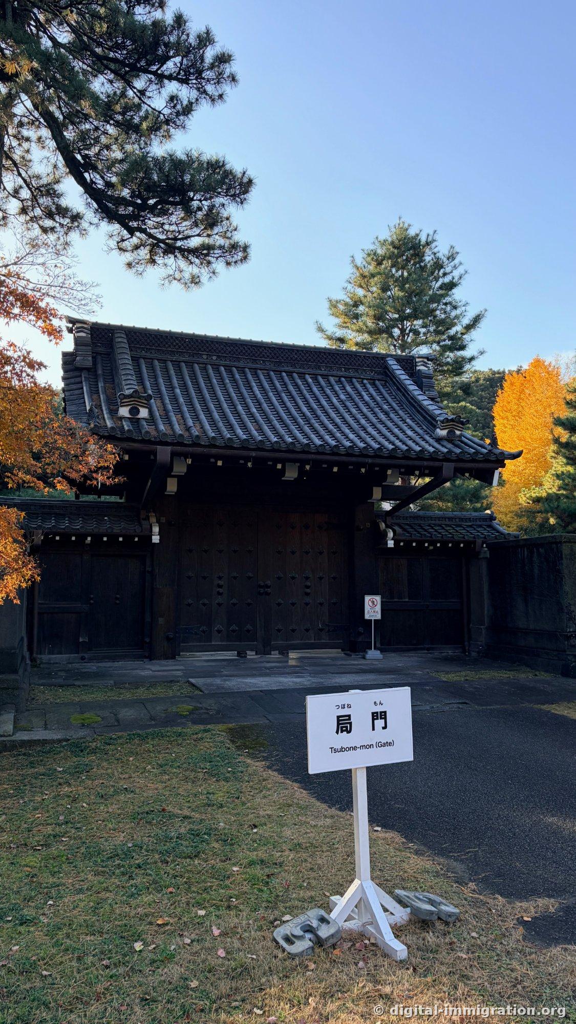 Devant une porte en bois noir se dresse un panneau blanc indiquant '局門 Tsubone-mon (Gate)' avec de l'érable et un ginkgo autour