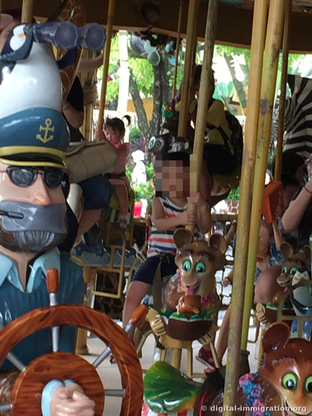 A child sitting in a lemur-shaped seat on the Madagascar character carousel, gripping the pole, with a penguin sculpture in a captain's hat