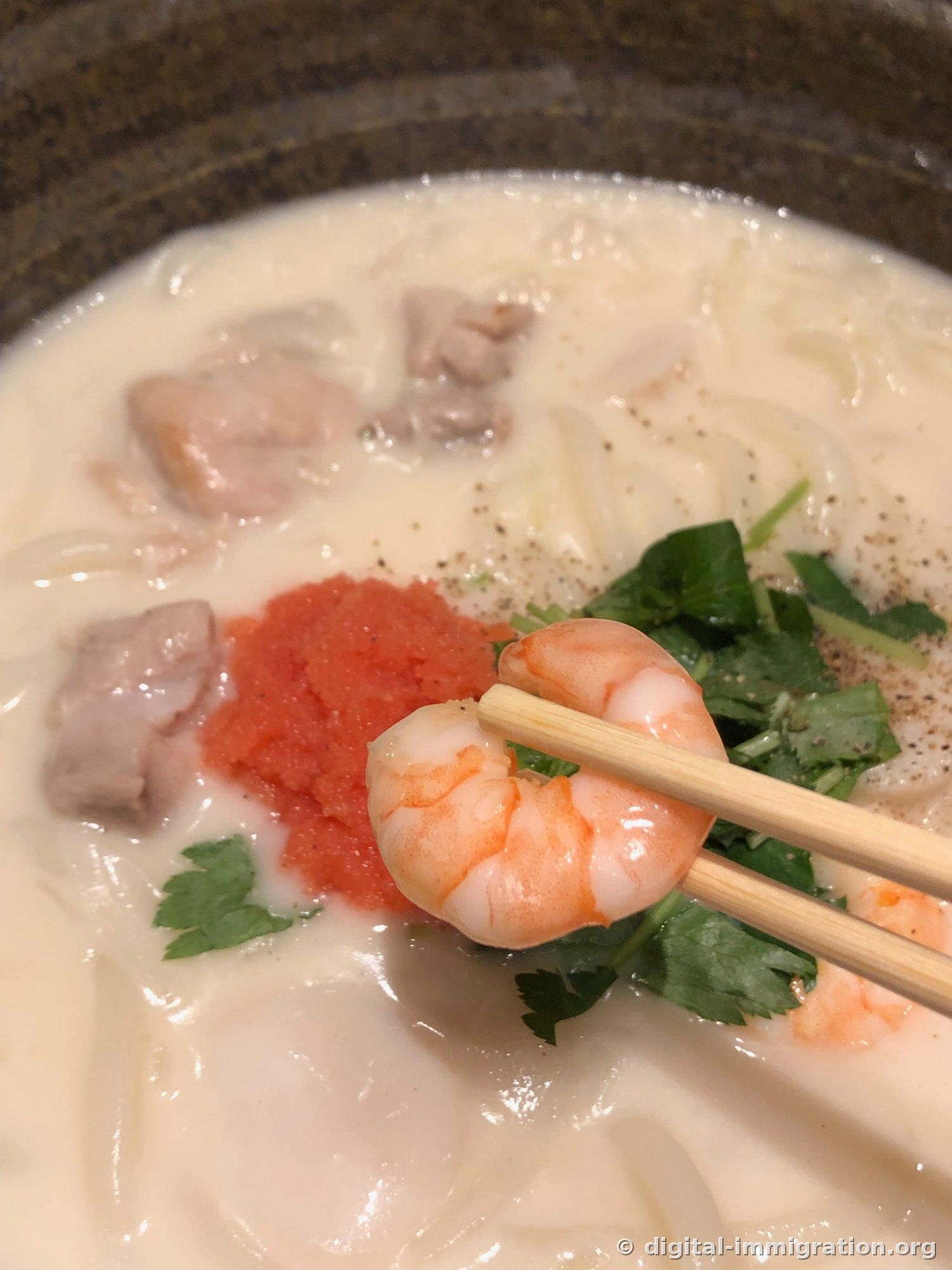 Close-up of a whole shrimp lifted from the cream broth with wooden chopsticks