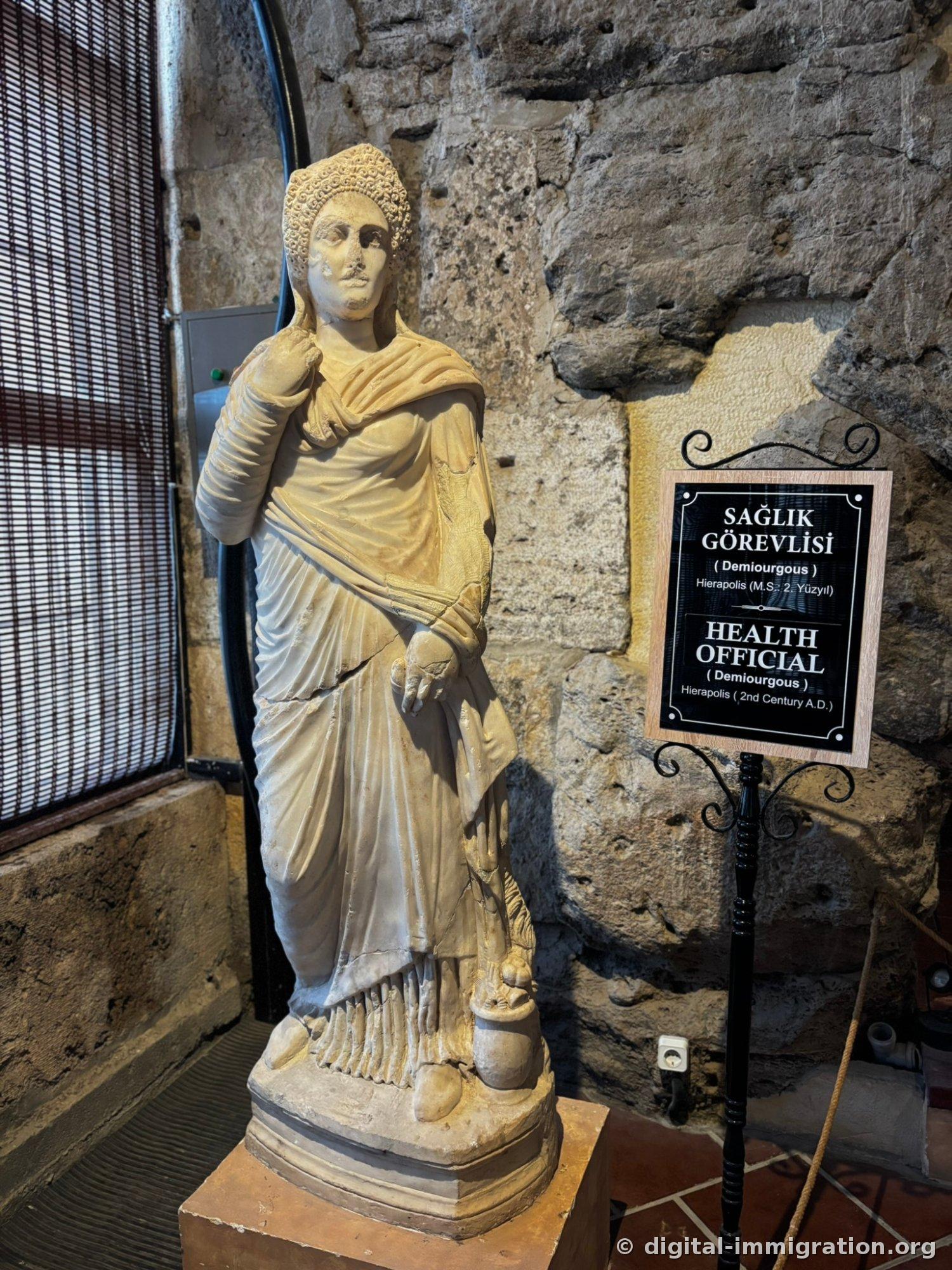 Full-length male statue wearing intricately carved curled-hair crown and toga, placard reading 'SAGLIK GOREVLISI / HEALTH OFFICIAL' beside it
