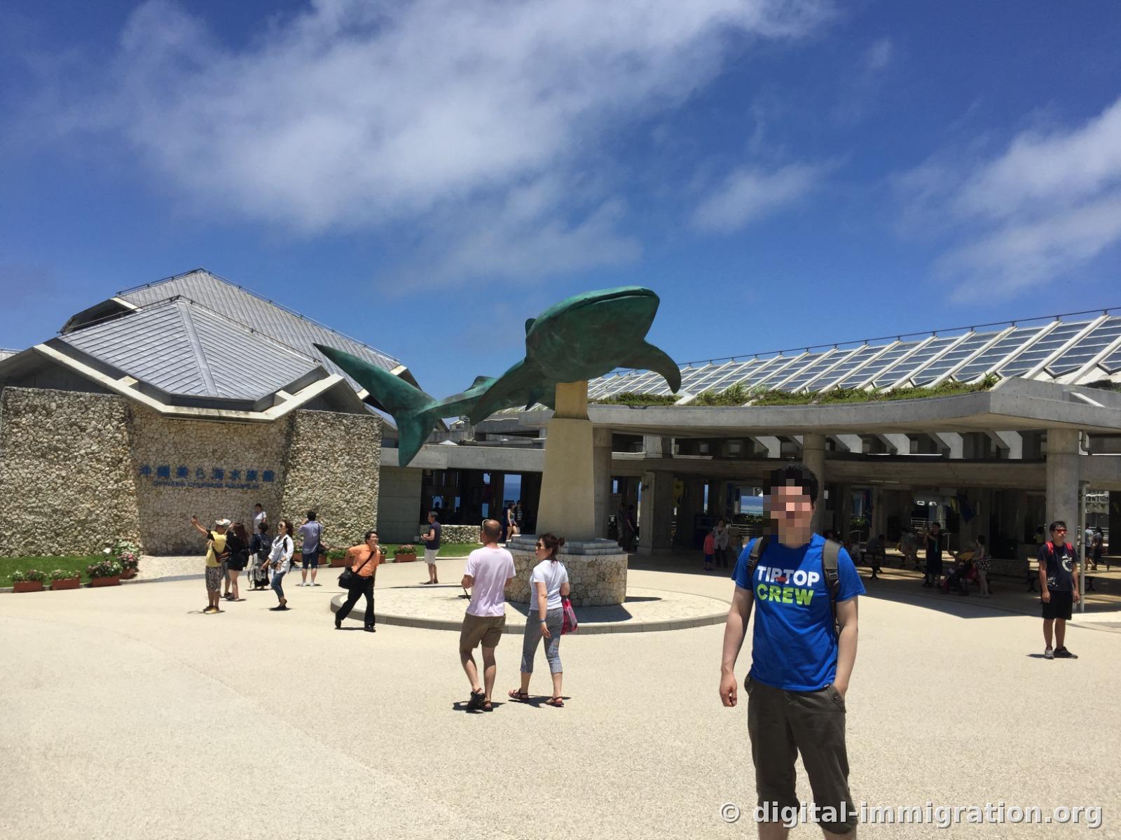 蓝色天空下美丽海水族馆入口广场，青铜鲸鲨雕像和玻璃屋顶主馆建筑，穿着T恤的游客们