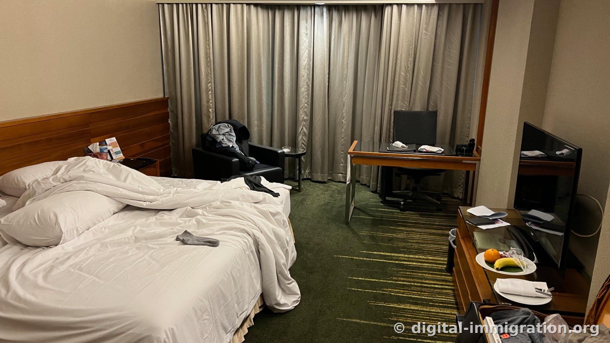 Chambre d'hôtel avec moquette verte, lit avec draps blancs, rideaux argentés, canapé noir, bureau en bois avec assiette de fruits sur la commode près de la TV