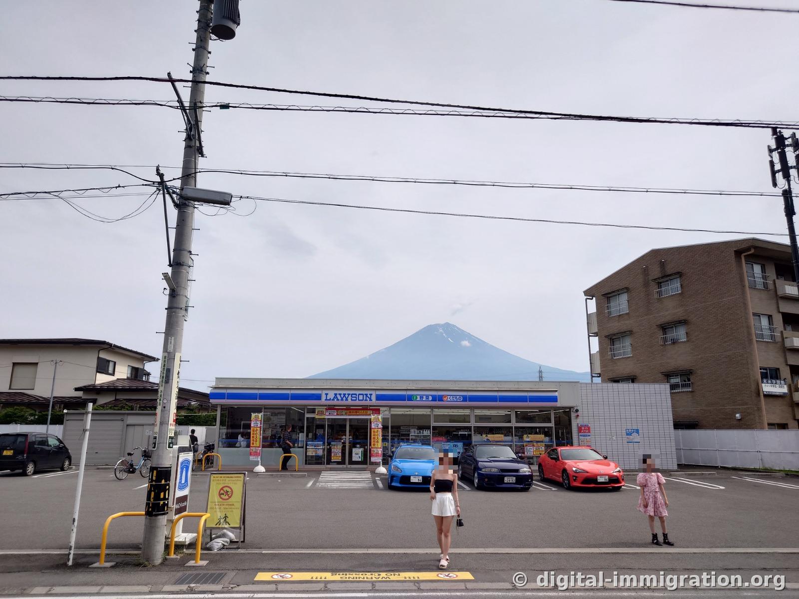 The Lawson storefront photographed from across the street with Mount Fuji rising above it, blue, navy, and red sports cars lined up in the parking lot, and tourists standing in front of the road