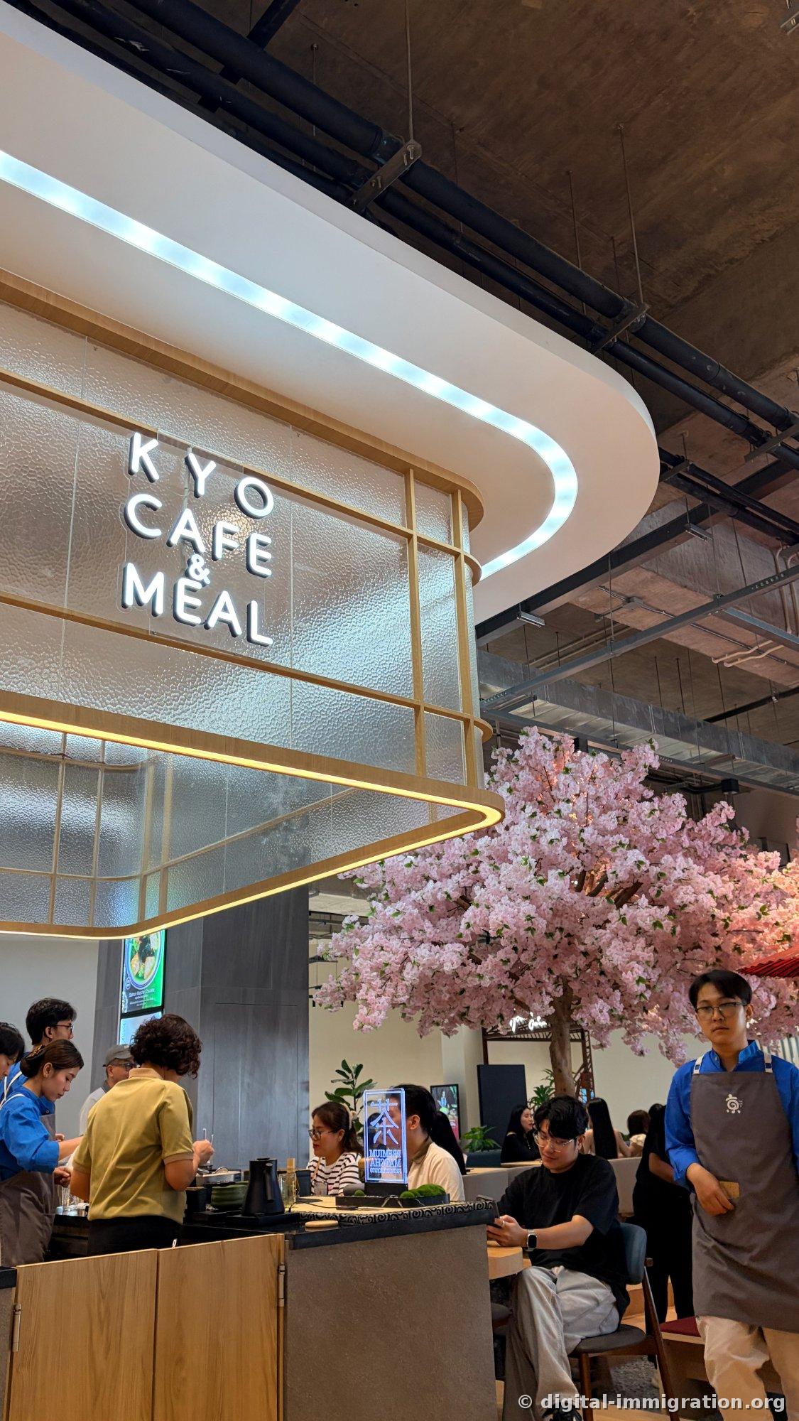 Inside view of the café with a white curved KYO CAFE & MEAL sign illuminated overhead against an exposed concrete ceiling, with a large pink artificial cherry tree, staff member in an apron, and seated customers visible
