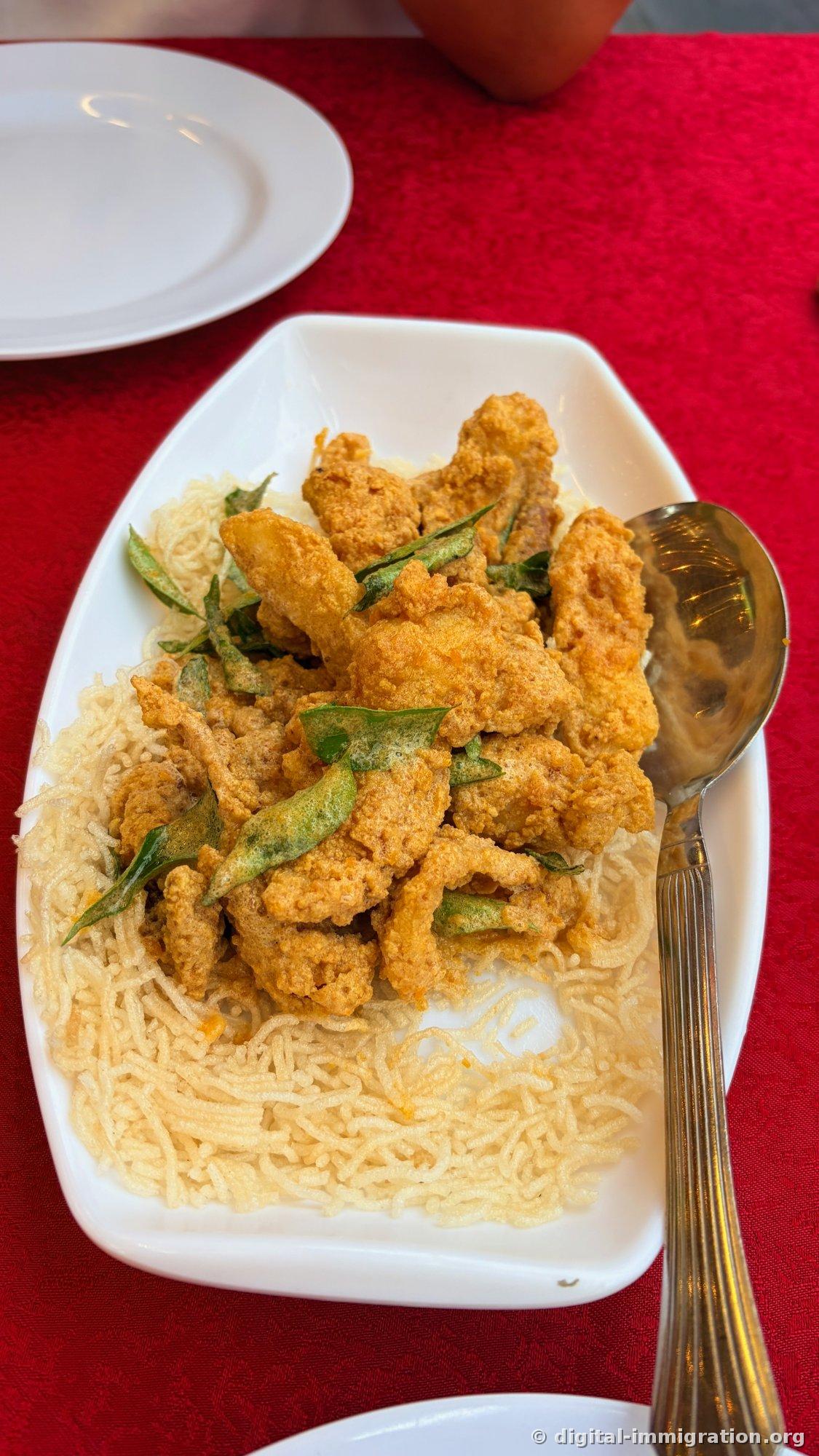 カリカリの揚げ麺の上にカレーバター・ソースでコーティングされた揚げた破片とカレーの揚げた葉が乗った料理が、白いお皿に盛られている
