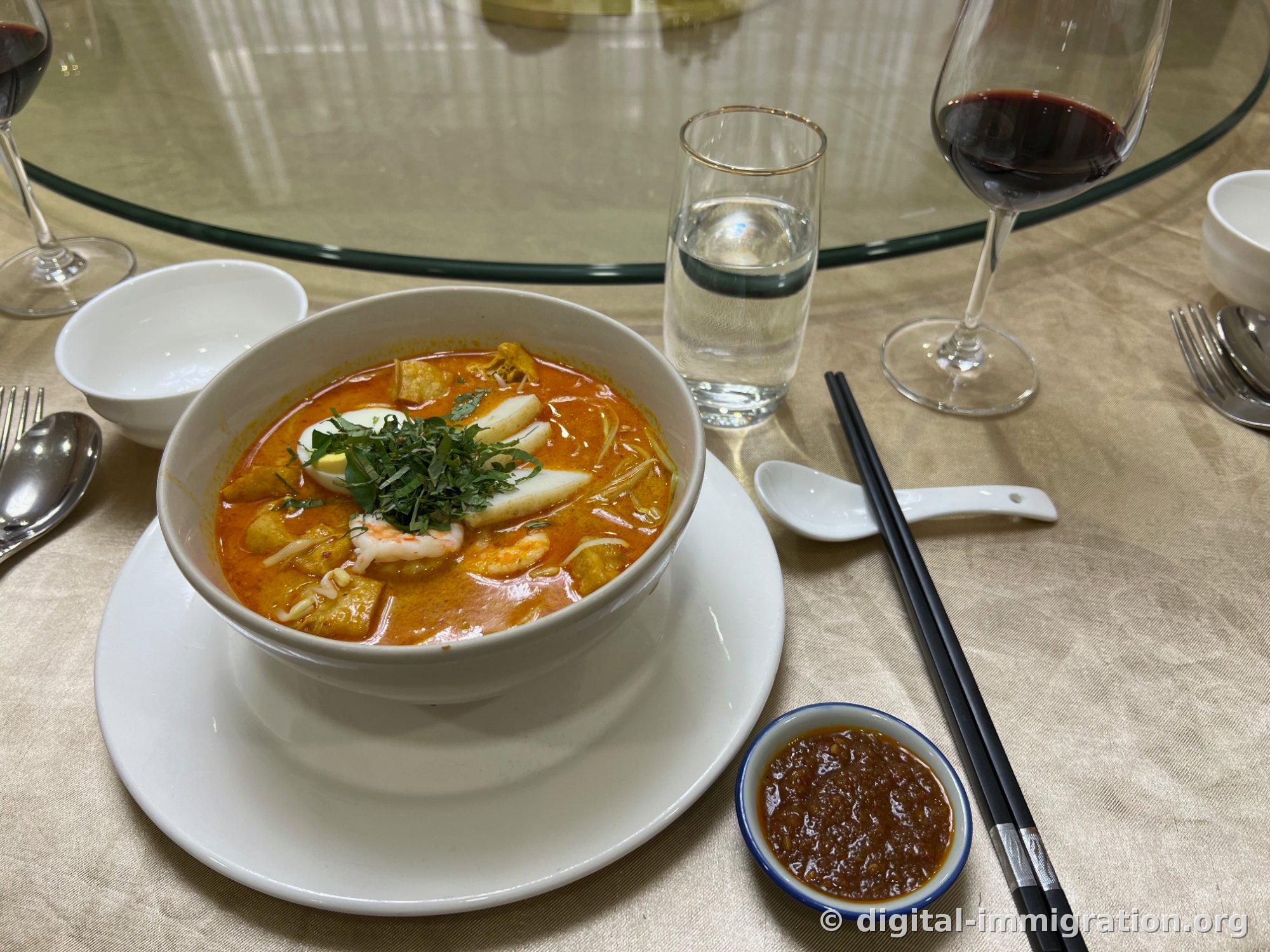 Laksa in a white bowl with orange coconut broth topped with boiled egg, fish cake, shrimp, bean sprouts, and minced laksa leaves, with a small dish of sambal and black chopsticks beside it