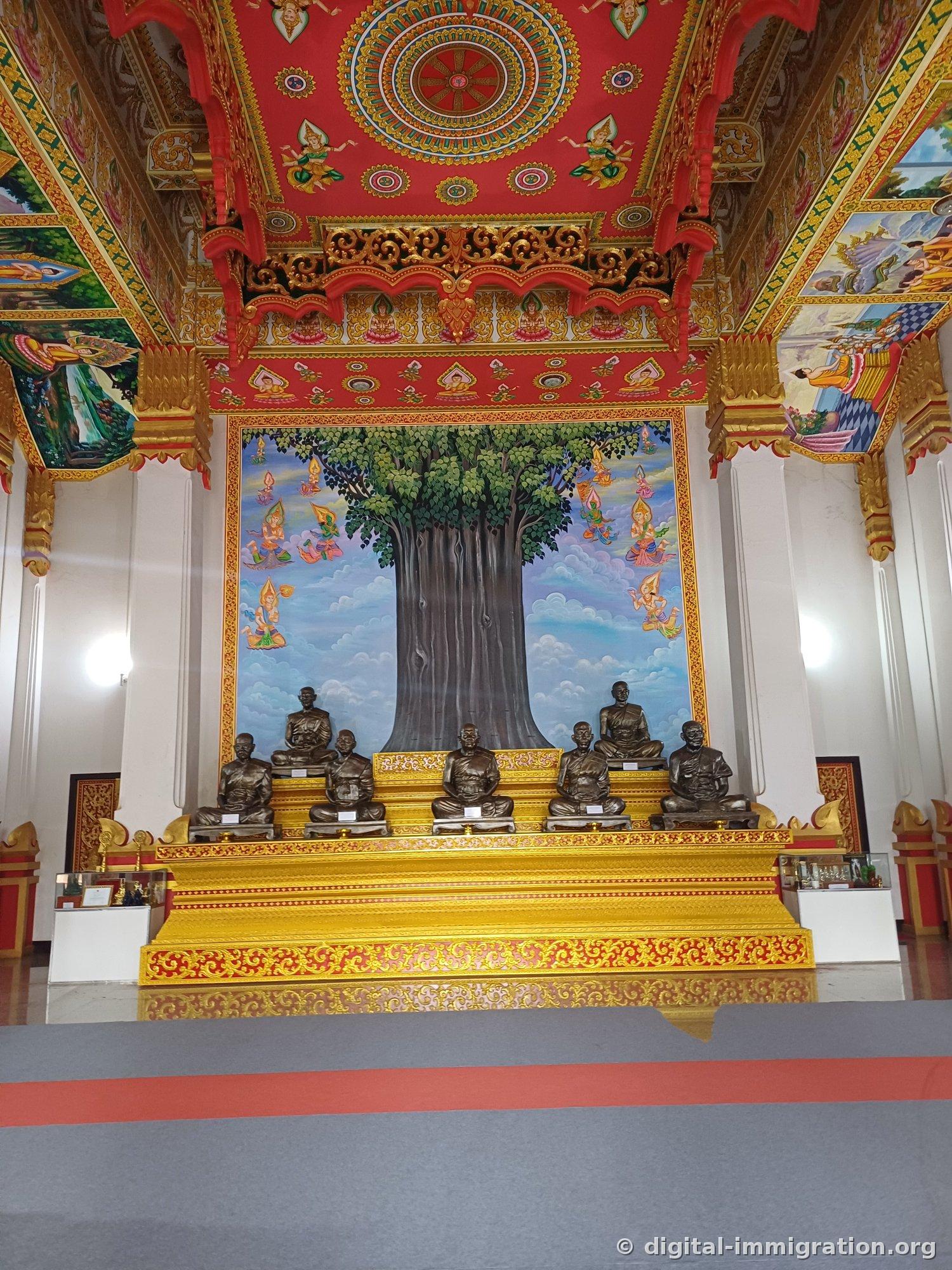 Interior of the ordination hall with Dhamma wheel pattern on red ceiling and six stone monk statues seated before Bodhi tree mural
