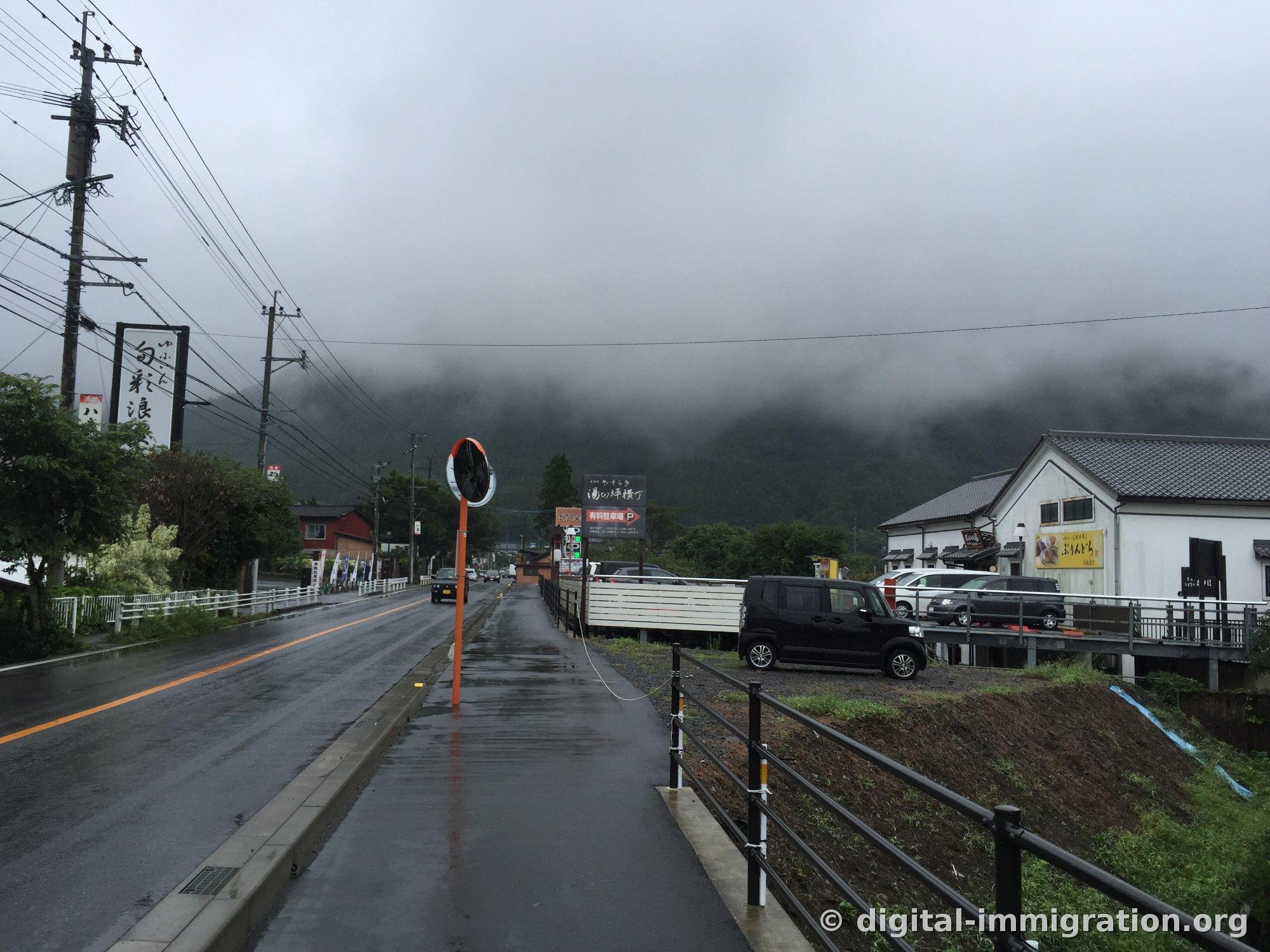 Une route mouillée par la pluie, un panneau de parking payant lisant '湯の坪横丁', une montagne basse enveloppée de nuages basse à Yufuin