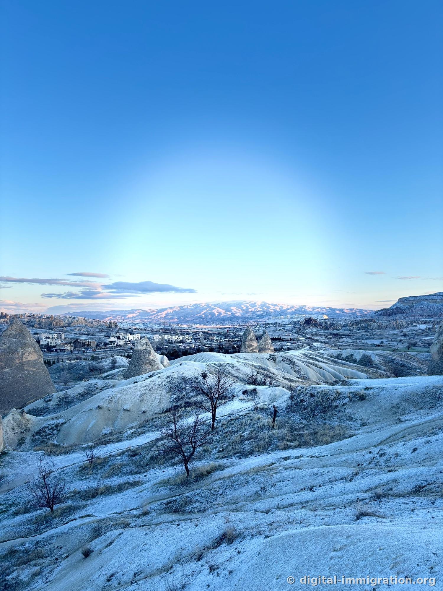 Juste avant l'aube, une lumière bleue douce, une colline de tuf couverte de givre et des arbres nus, au loin les bâtiments blancs du village de Göreme et une crête de montagne enneigée tintée de rose
