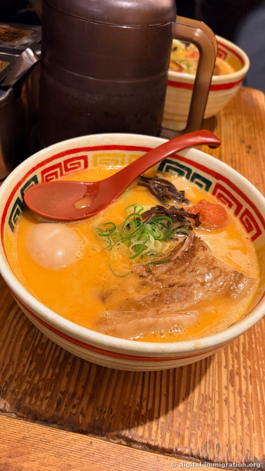 A bowl of ramen with orange-tinted tonkotsu broth topped with ajitsuke tamago, thinly sliced scallion, kikurage mushroom, a whole lump of mentaiko, and a piece of kakuni with visible grain, with a red renge spoon