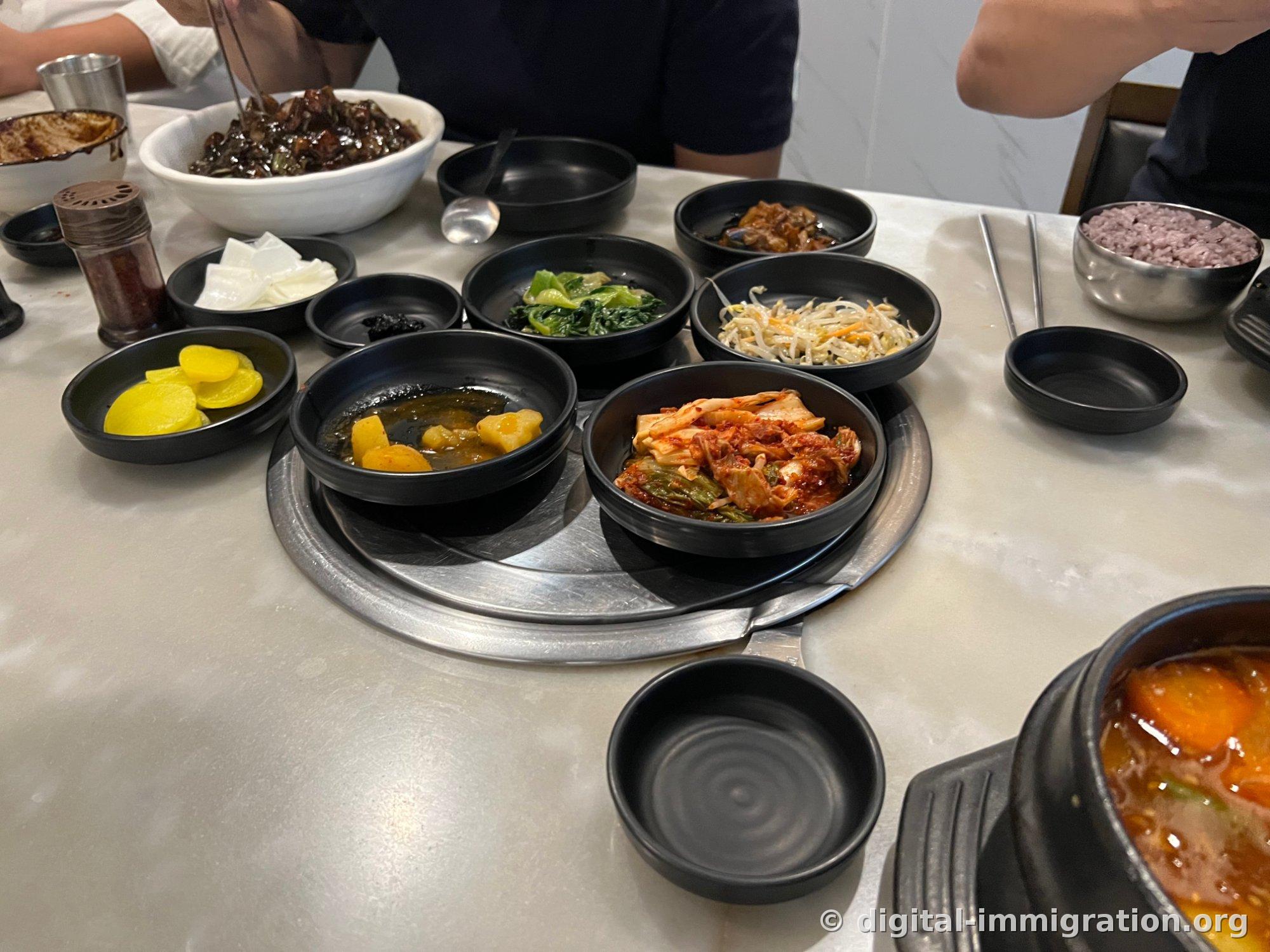 Bols noirs de banchan sur une table en marbre blanc. Danmuji, radis mariné, épinards et pak-choï assaisonnés, pousses de soja assaisonnées, kimchi de chou chinois et pommes de terre mijotées disposés sur un plateau tournant, avec en arrière-plan un bol de jjajangmyeon et du riz noir.