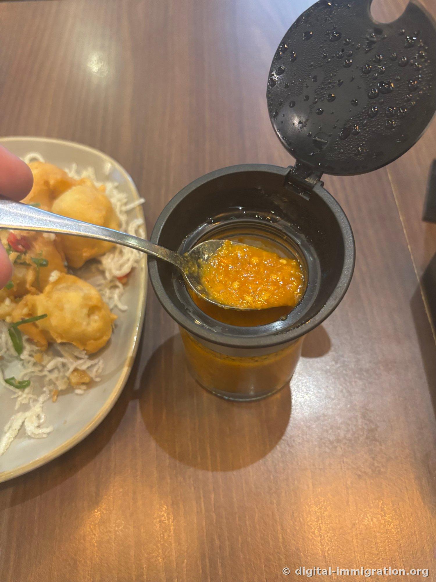 Spooning orange-colored sambal from an open glass bottle with black cap, with the fried soft tofu plate on the left