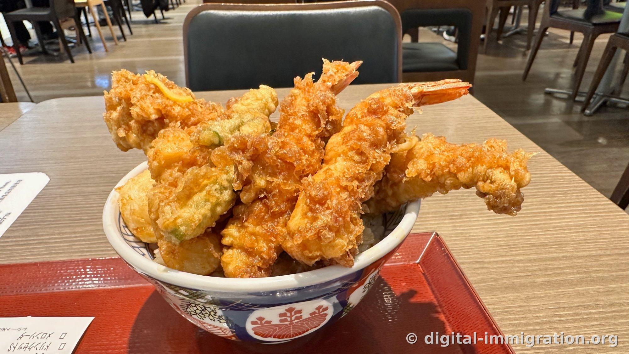 A fried shrimp donburi piled so high the rice bowl is hidden, two large shrimp, vegetables like pumpkin tempura, and a long eel tempura extending across the edge of the ceramic bowl on a food court table